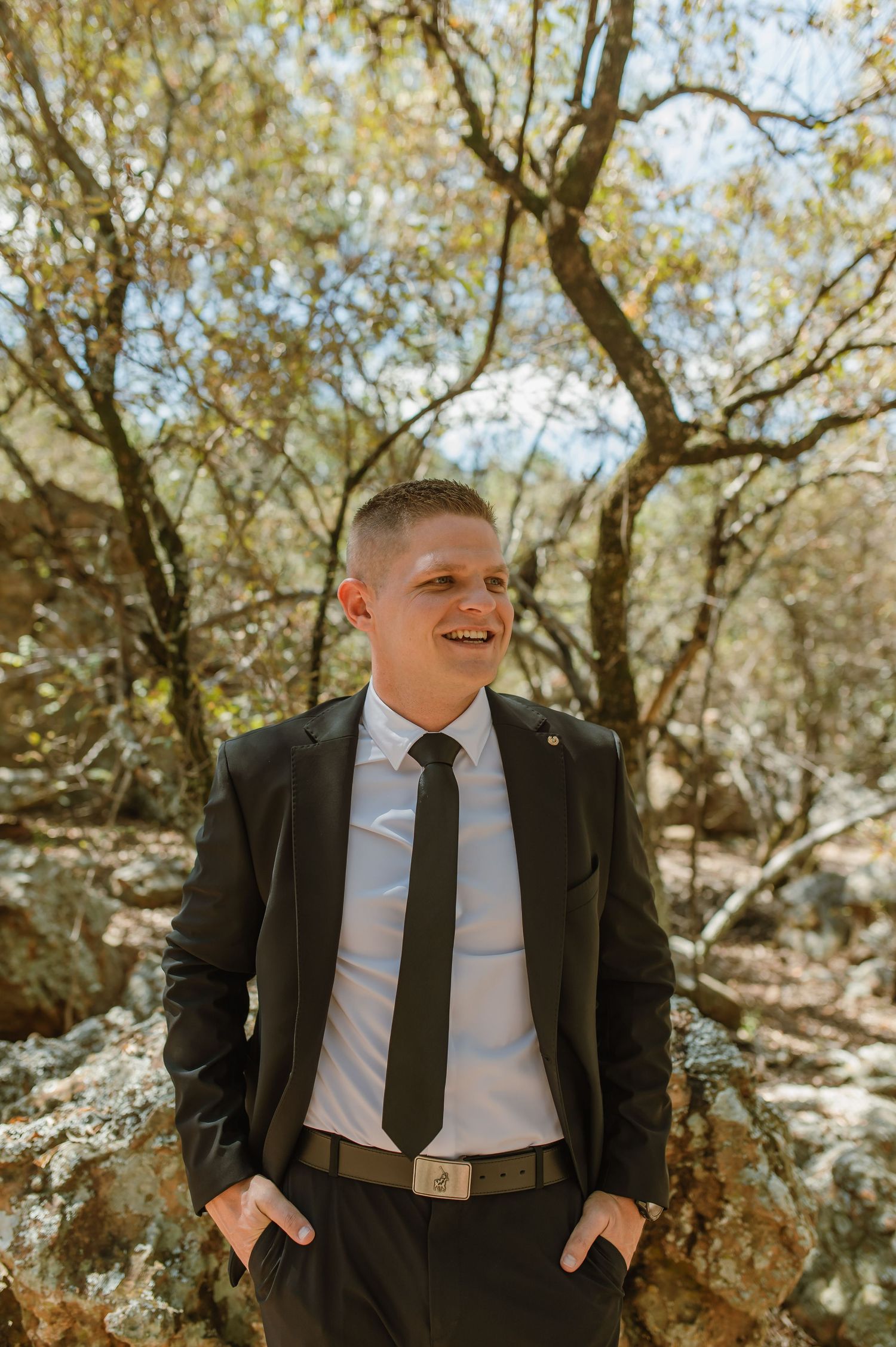 Smiling groom in dark suit and black tie standing outdoors among rocky terrain and trees.