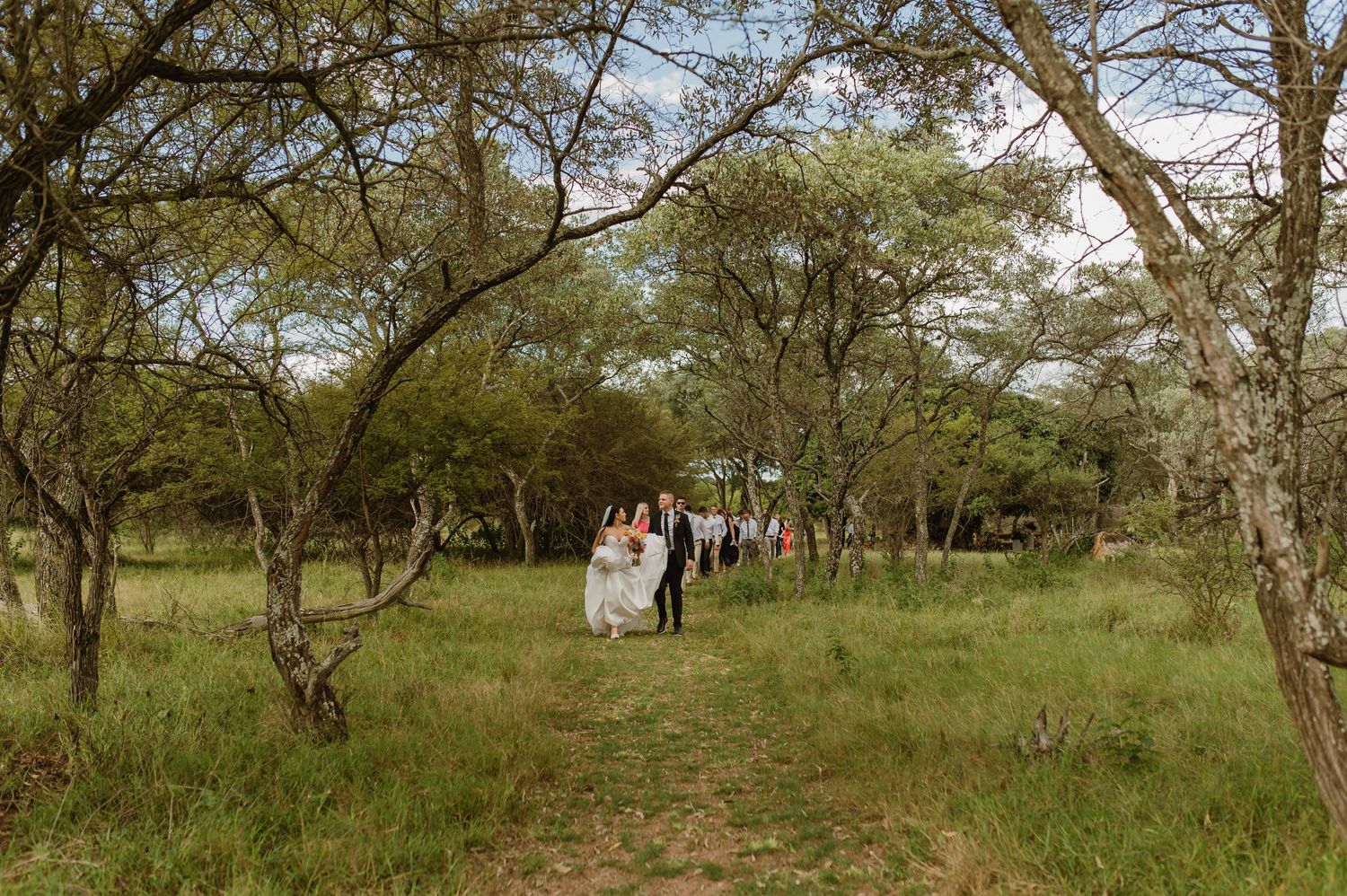 Wedding party walks along a dirt path through lush green trees toward outdoor ceremony.