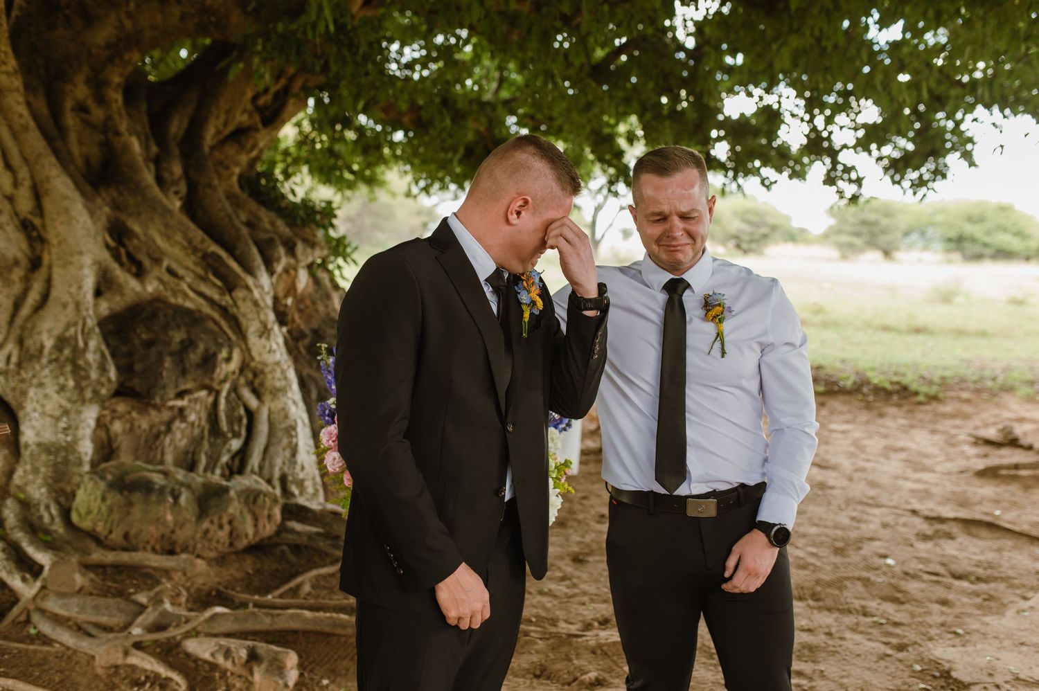 Emotional groom in a dark suit being comforted by his groomsman under a large tree before the ceremony.