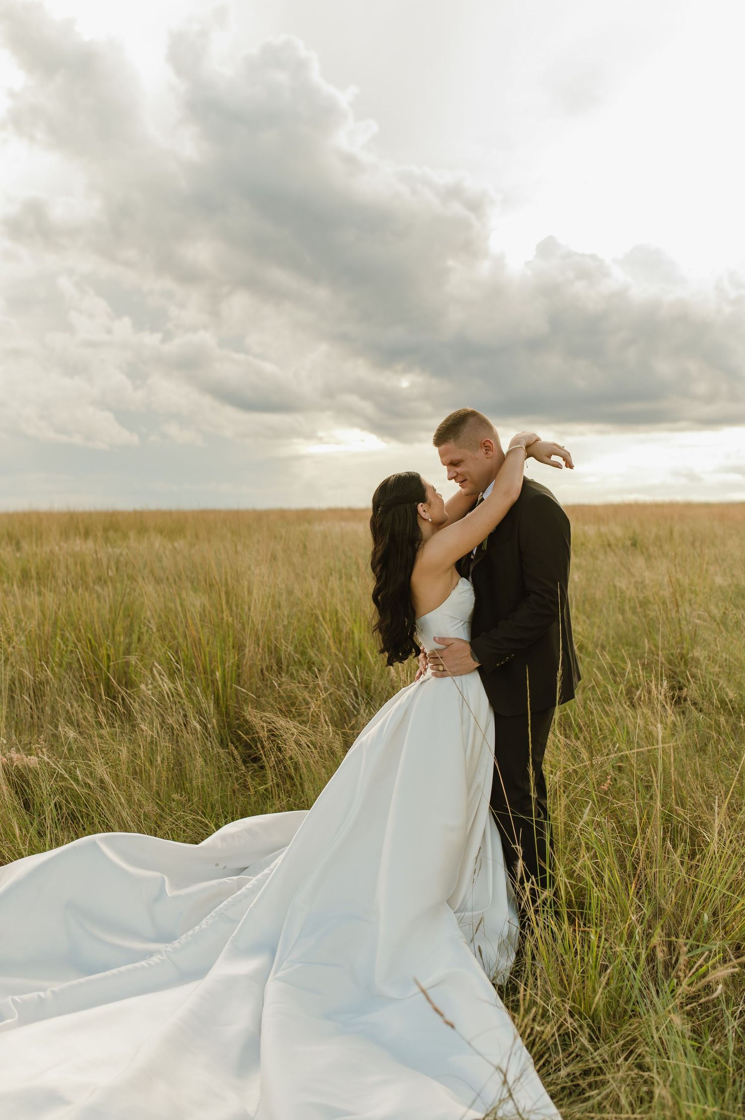 Bride and groom embrace romantically in golden grass field under dramatic cloudy sky.
