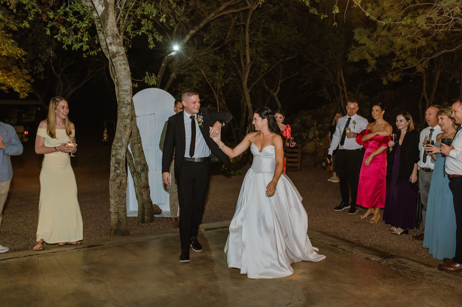 Bride and groom share first dance outdoors at night surrounded by wedding guests under string lights.
