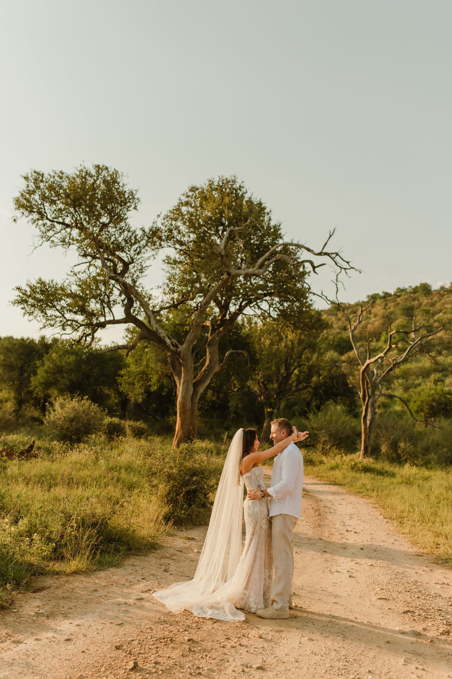 Bride and groom share romantic moment on sandy bush path at sunset, her long veil flowing in golden light.
