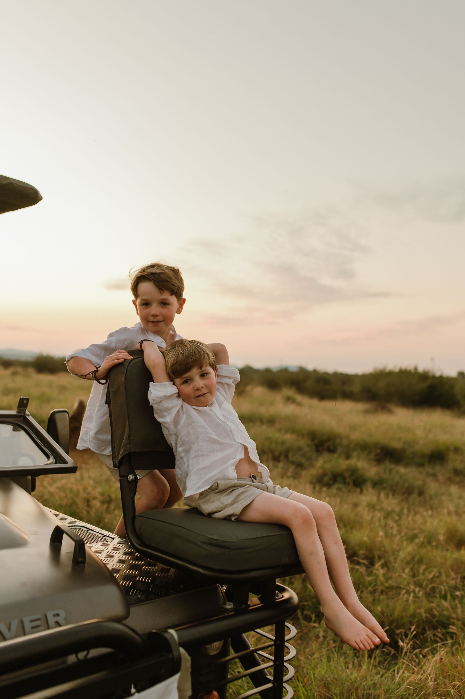 Two young boys sitting together on a Land Rover at sunset.