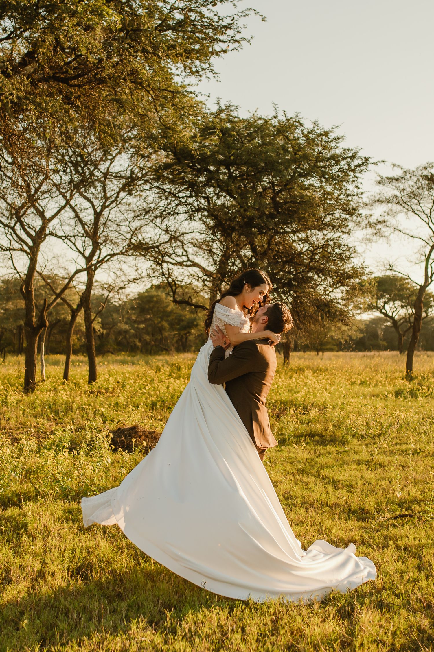 A romantic wedding couple dances in a sunlit field surrounded by trees during golden hour.