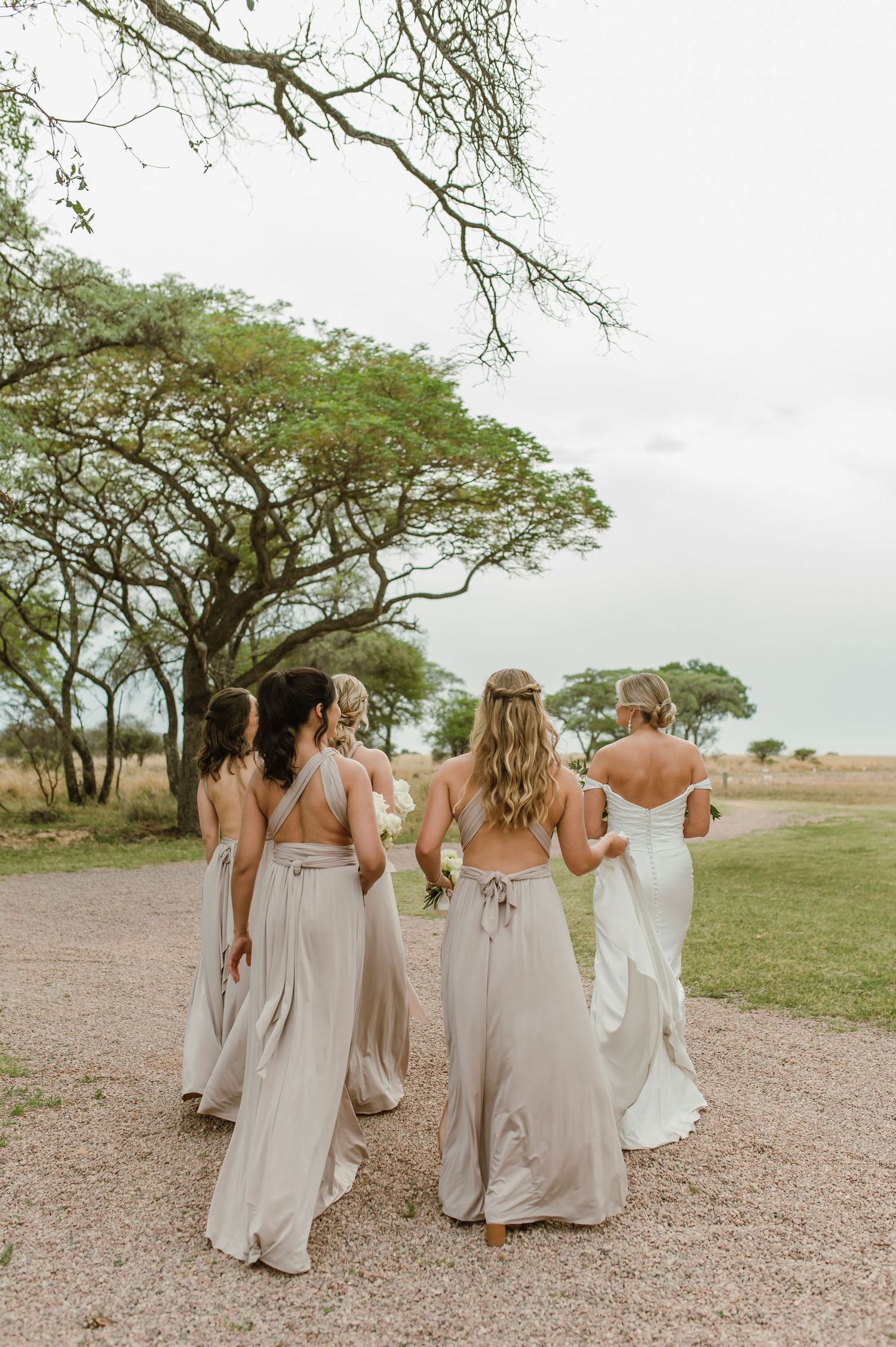 Bridesmaids in neutral dresses walking together on a gravel path surrounded by trees.