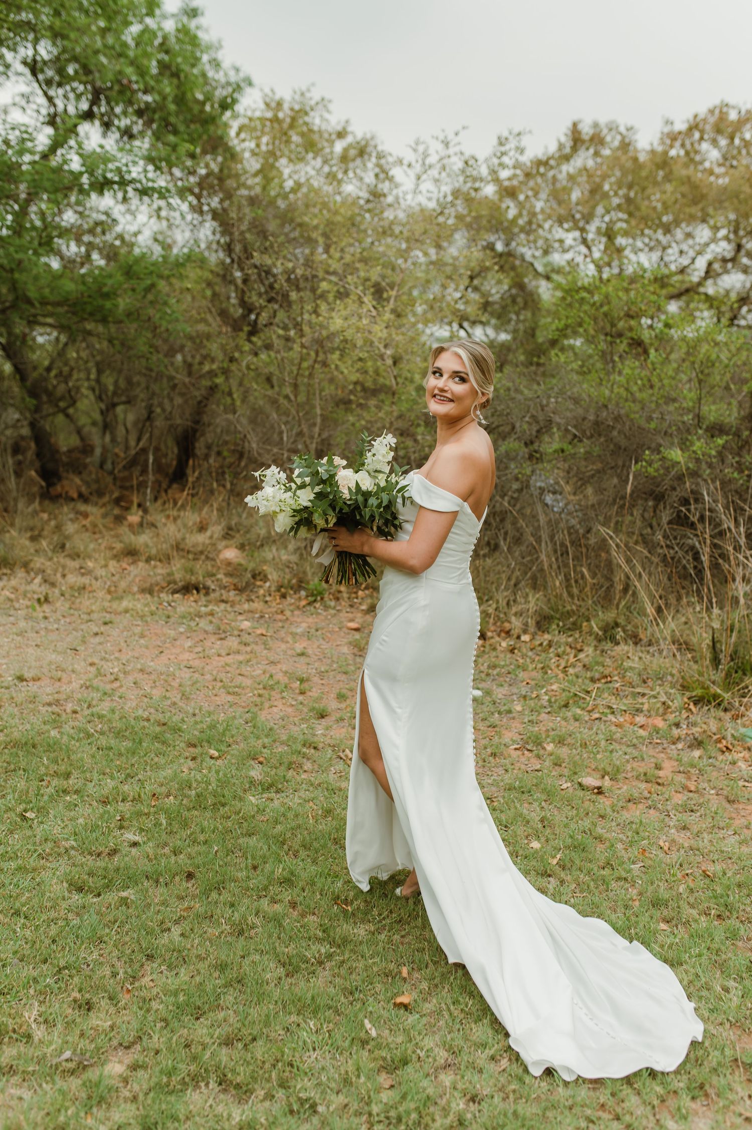 Elegant bride in off-shoulder gown holds white bouquet against natural landscape.