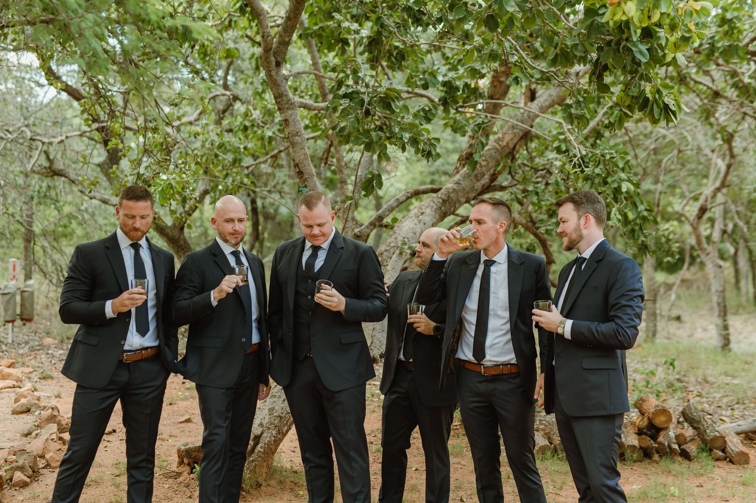 Wedding party in black suits posed casually among trees in natural lighting.