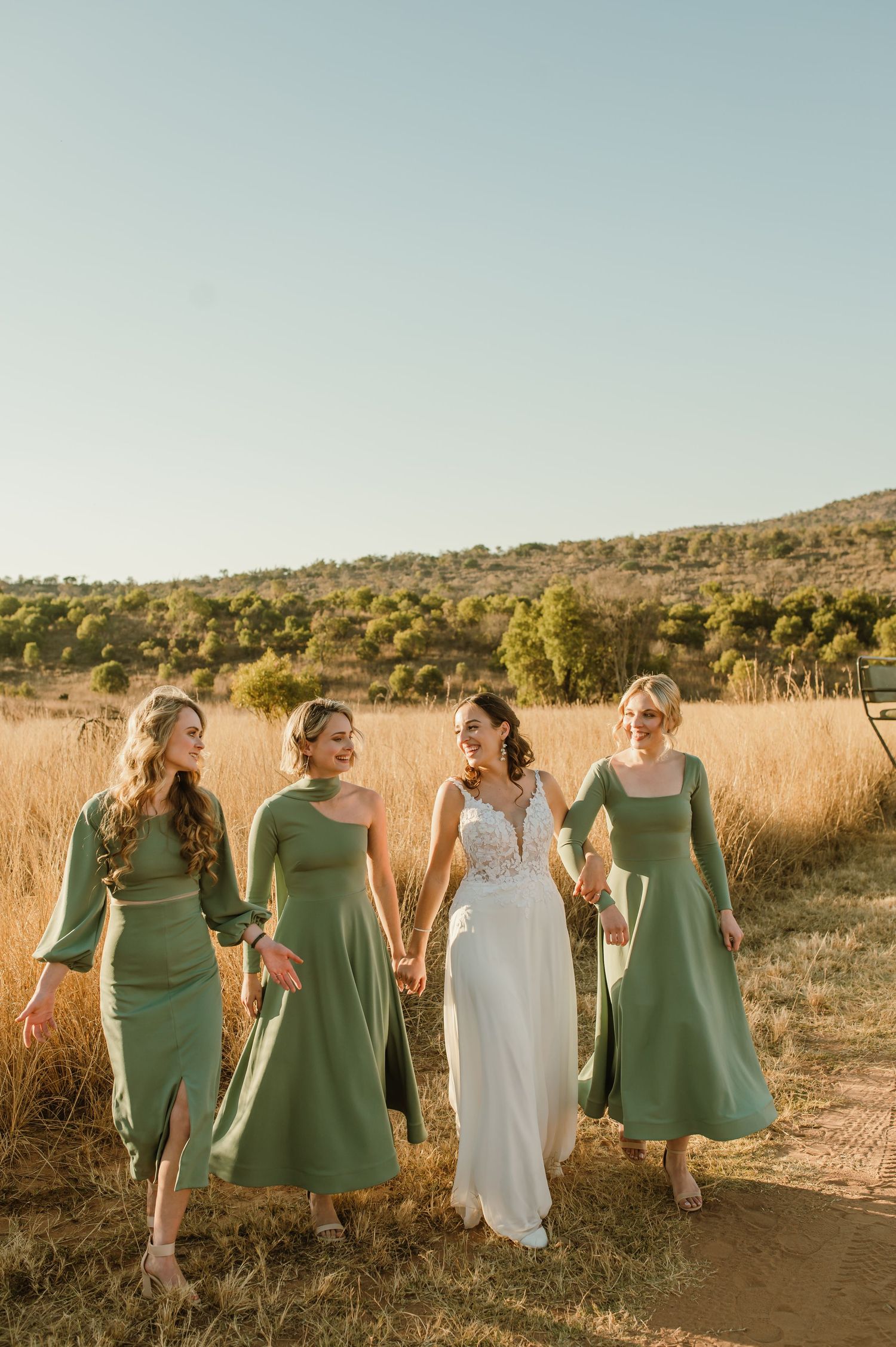Bridesmaids in sage green dresses walk through tall grass at golden hour.