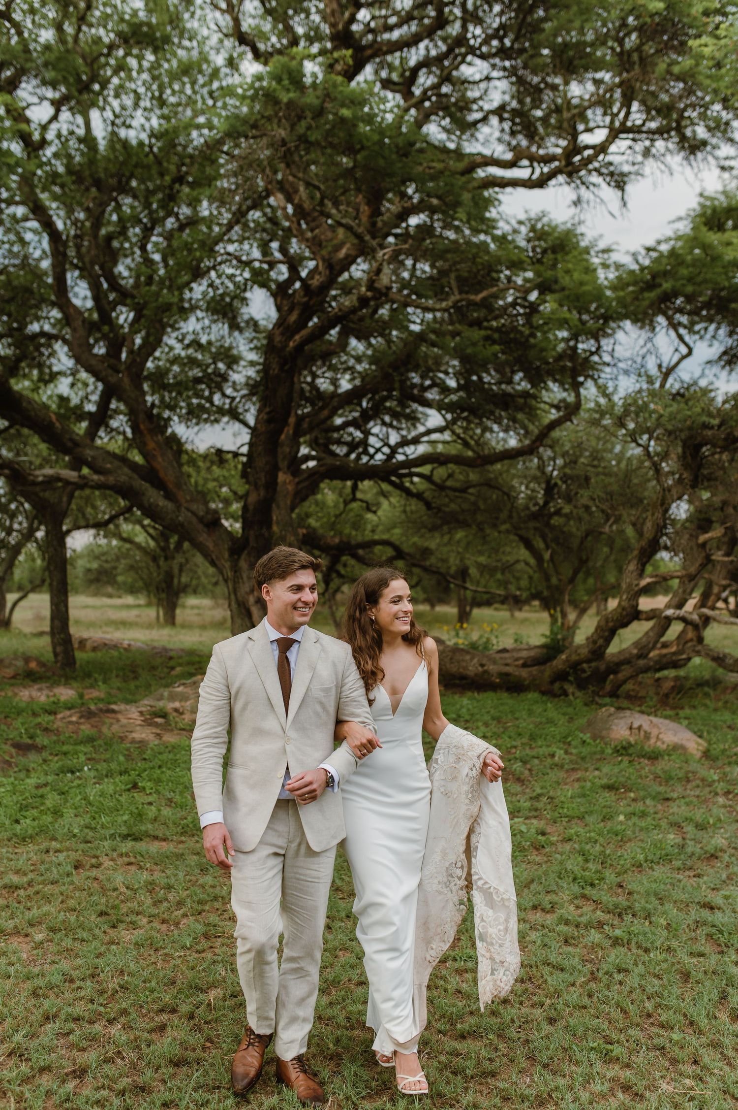 Newlyweds walking and laughing together through a scenic orchard.