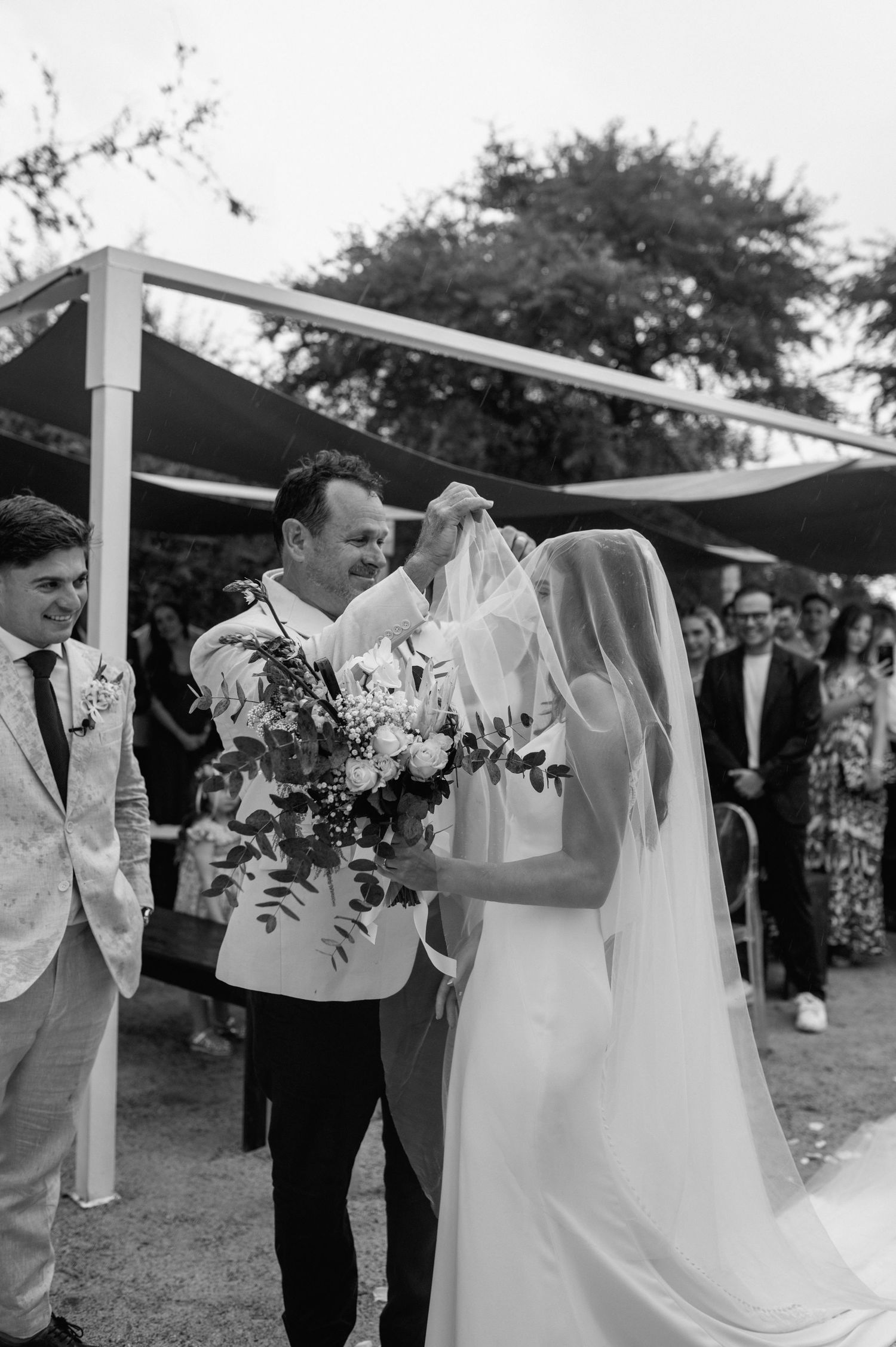 A black and white photo captures a wedding ceremony moment under an outdoor canopy structure.