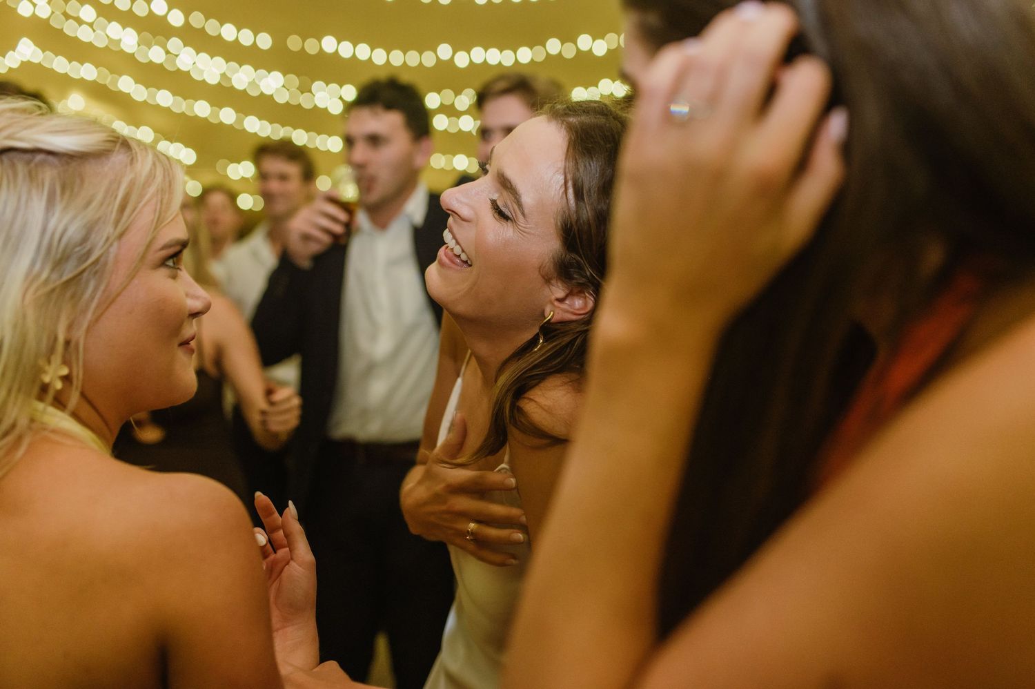Wedding reception guests dancing and celebrating under warm string lights at an indoor venue.