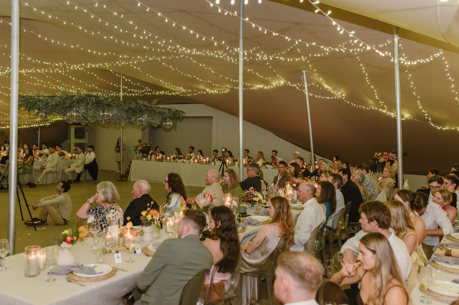 Panoramic view of wedding reception tent with guests seated at long tables under string lights.