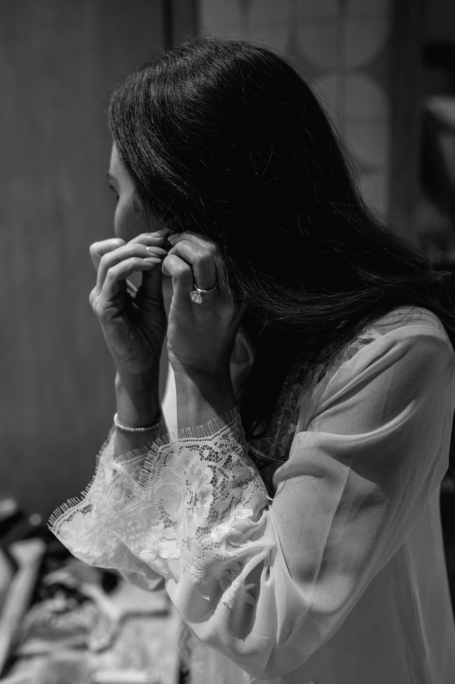 Black and white photo of bride in lace robe putting on earrings while getting ready for wedding.