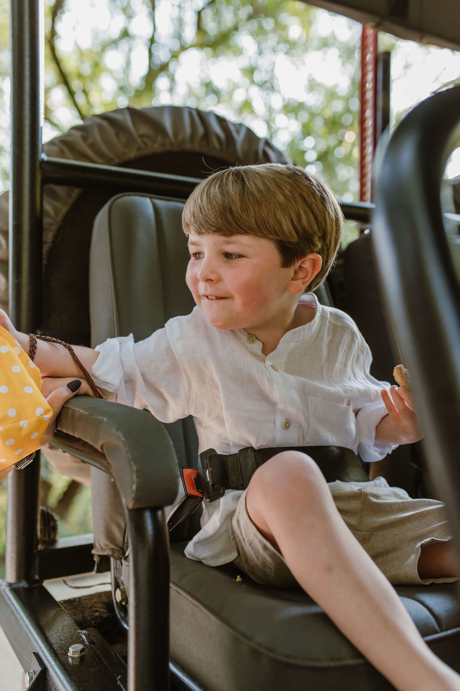 A happy young boy in a white linen shirt sits buckled in a safari vehicle seat.