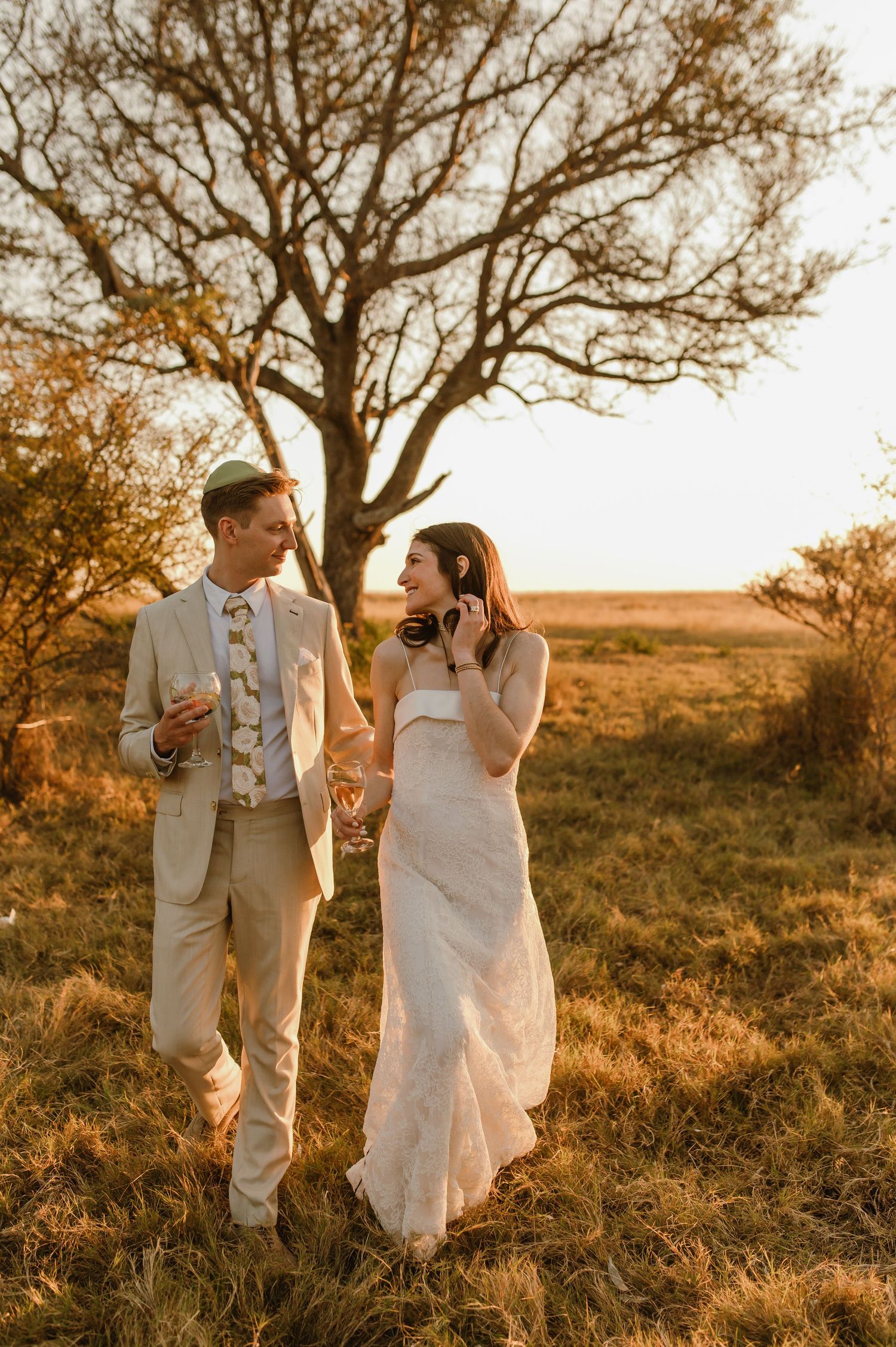 Bride and groom walk through golden field at sunset, she holds headphones, he holds champagne, bare tree backdrop.