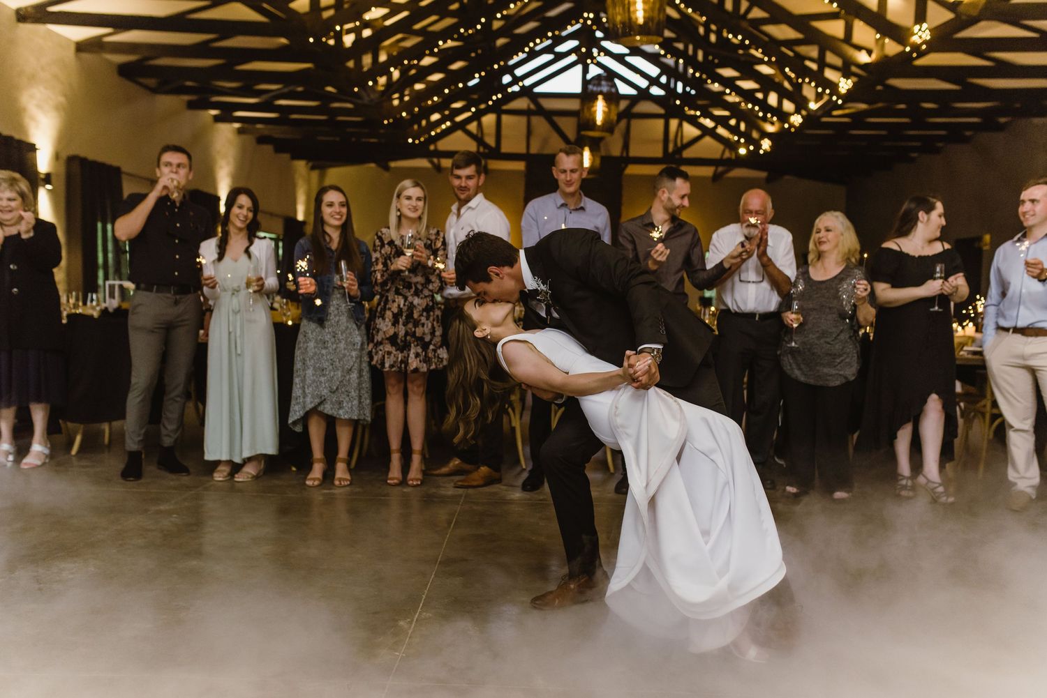 Wedding guests watch as dancers perform on a misty dance floor under rustic wooden beams.
