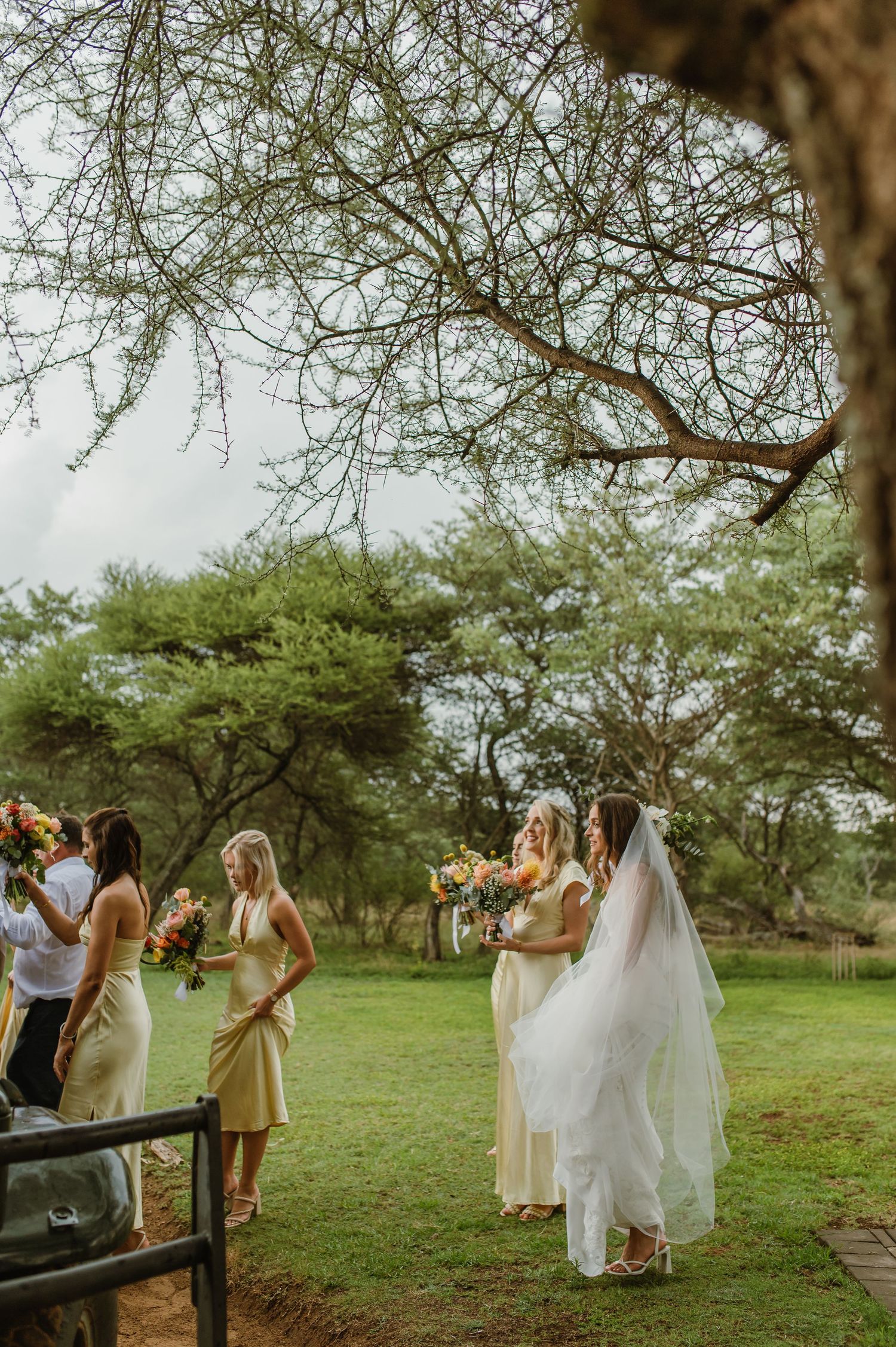 Wedding party gathered outdoors under acacia trees holding vibrant bouquets.