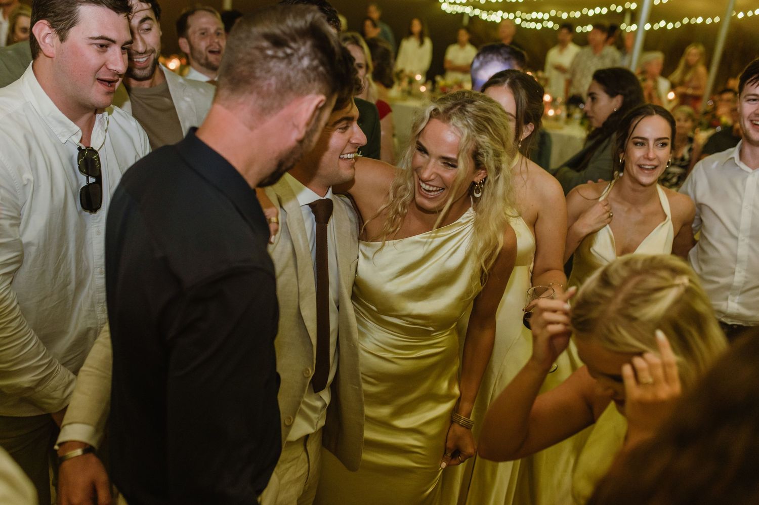 Wedding party celebrates with energetic dancing under golden string lights.