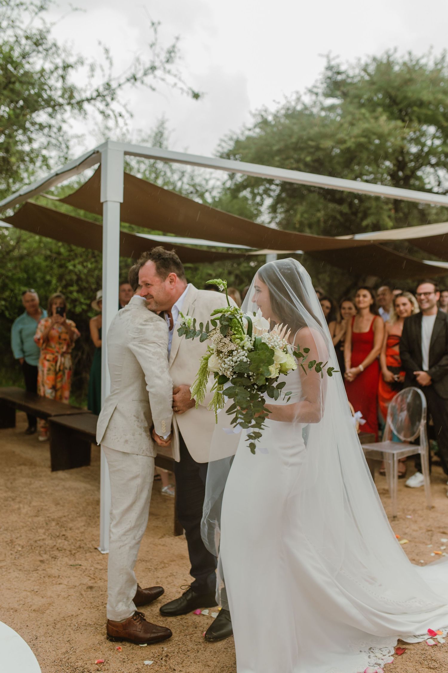 An outdoor wedding ceremony takes place under brown canopies with guests watching from the sides.
