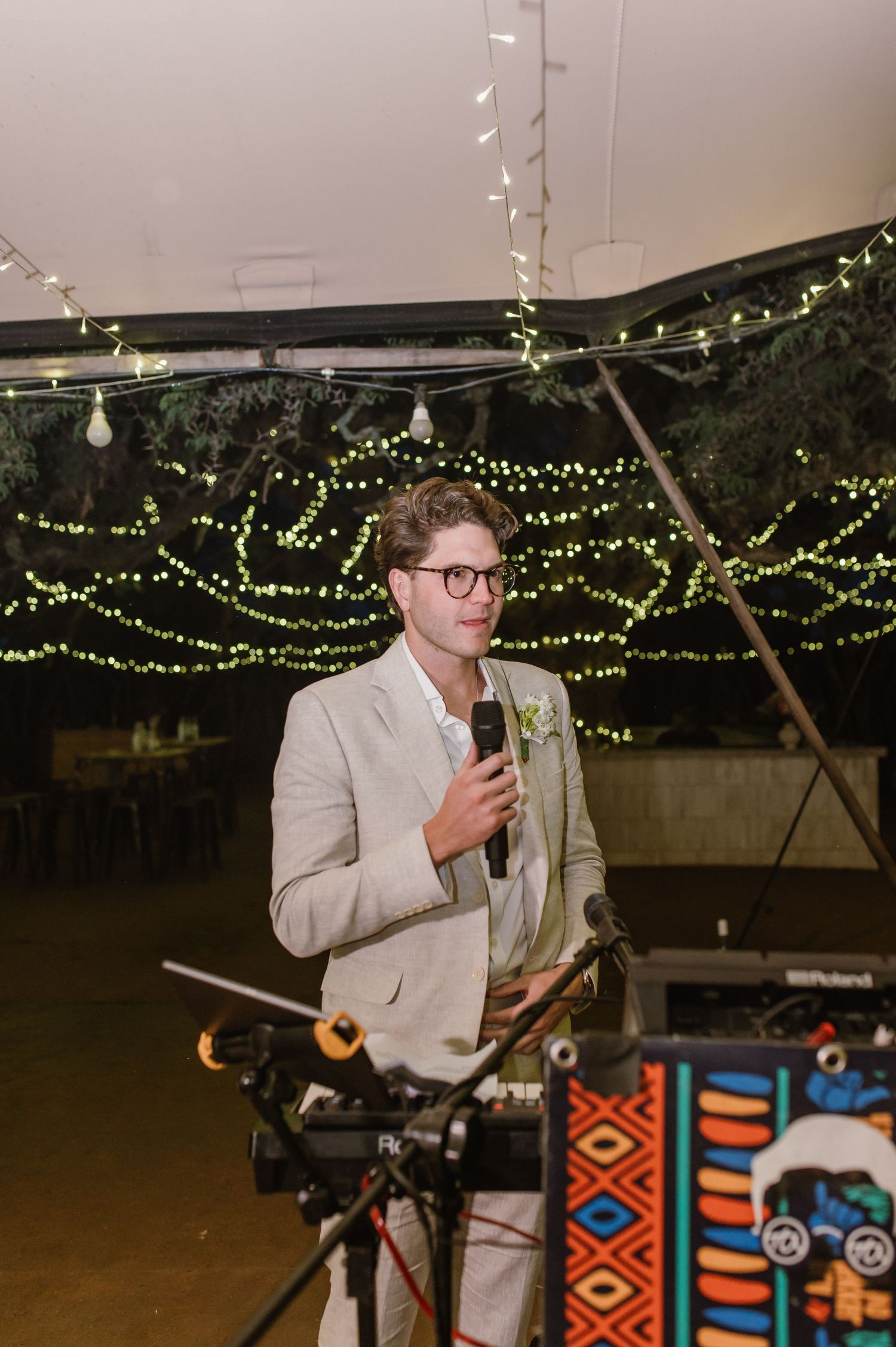 Speaker at wedding reception standing at decorated podium under string lights.