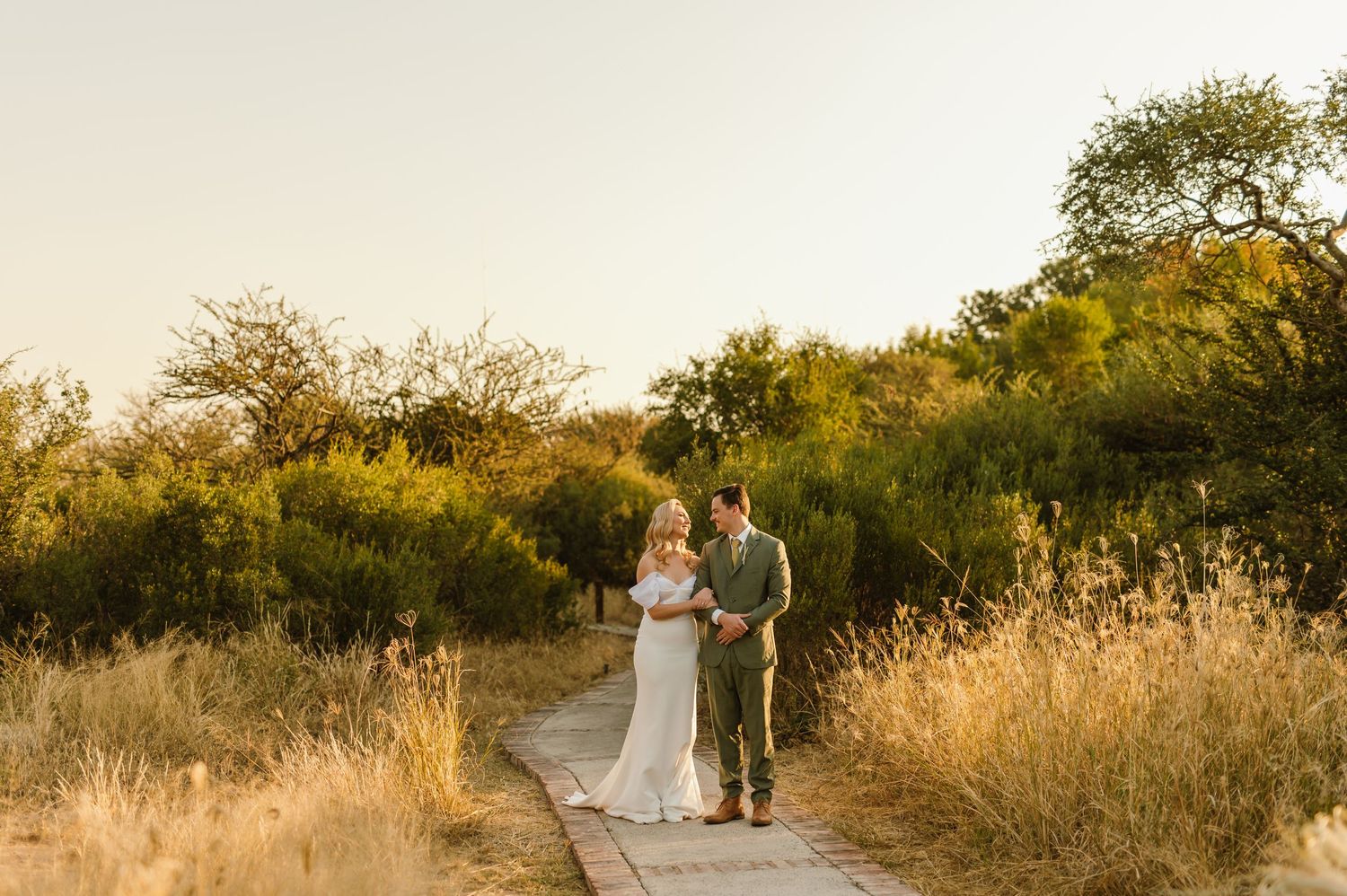 Wedding couple strolls down a winding path surrounded by desert brush at sunset.
