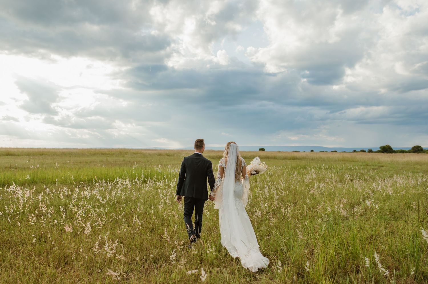 Wedding couple walking through a serene field with dramatic cloudy sky overhead.