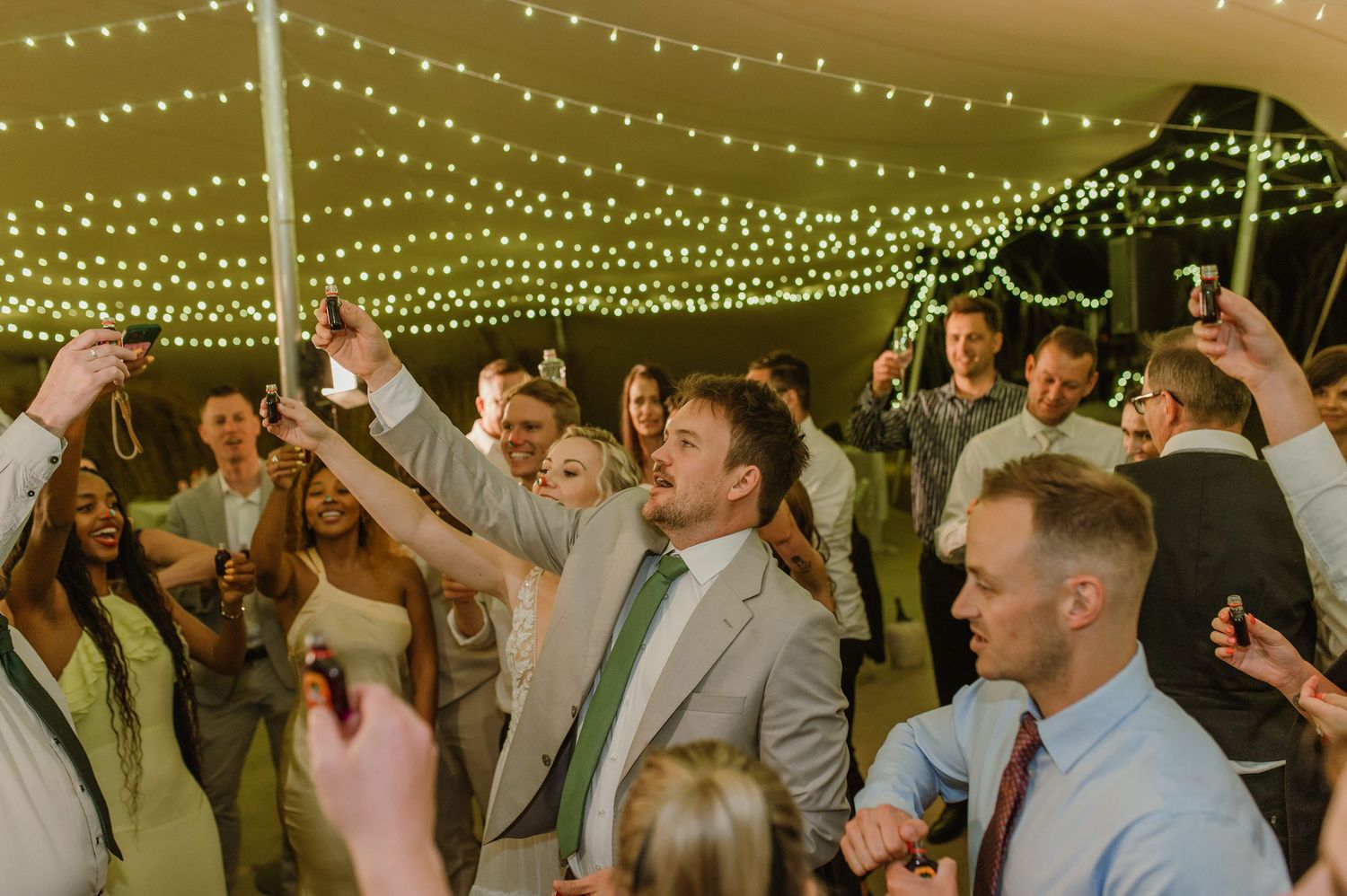 Wedding guests dancing and celebrating with raised arms under canopy of string lights.
