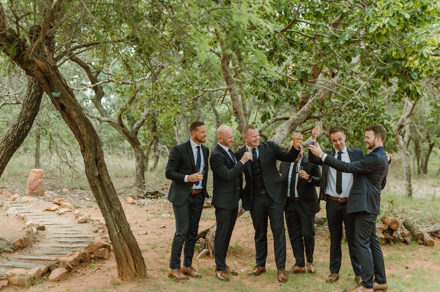 Groomsmen in formal attire gathered under trees for wedding photos.