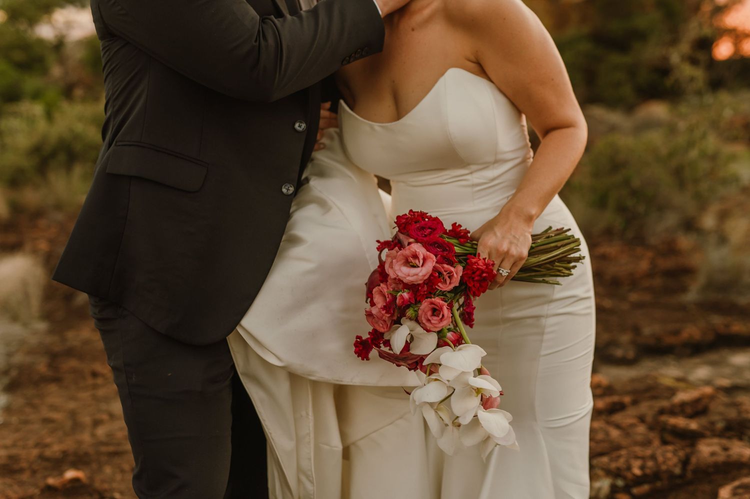 A couple embraces during their wedding photoshoot with red floral bouquet details.