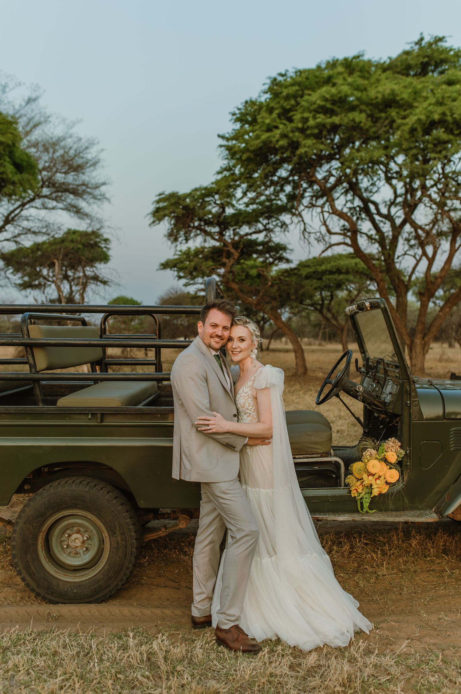 Elegant wedding couple embraces next to vintage safari vehicle in African landscape.