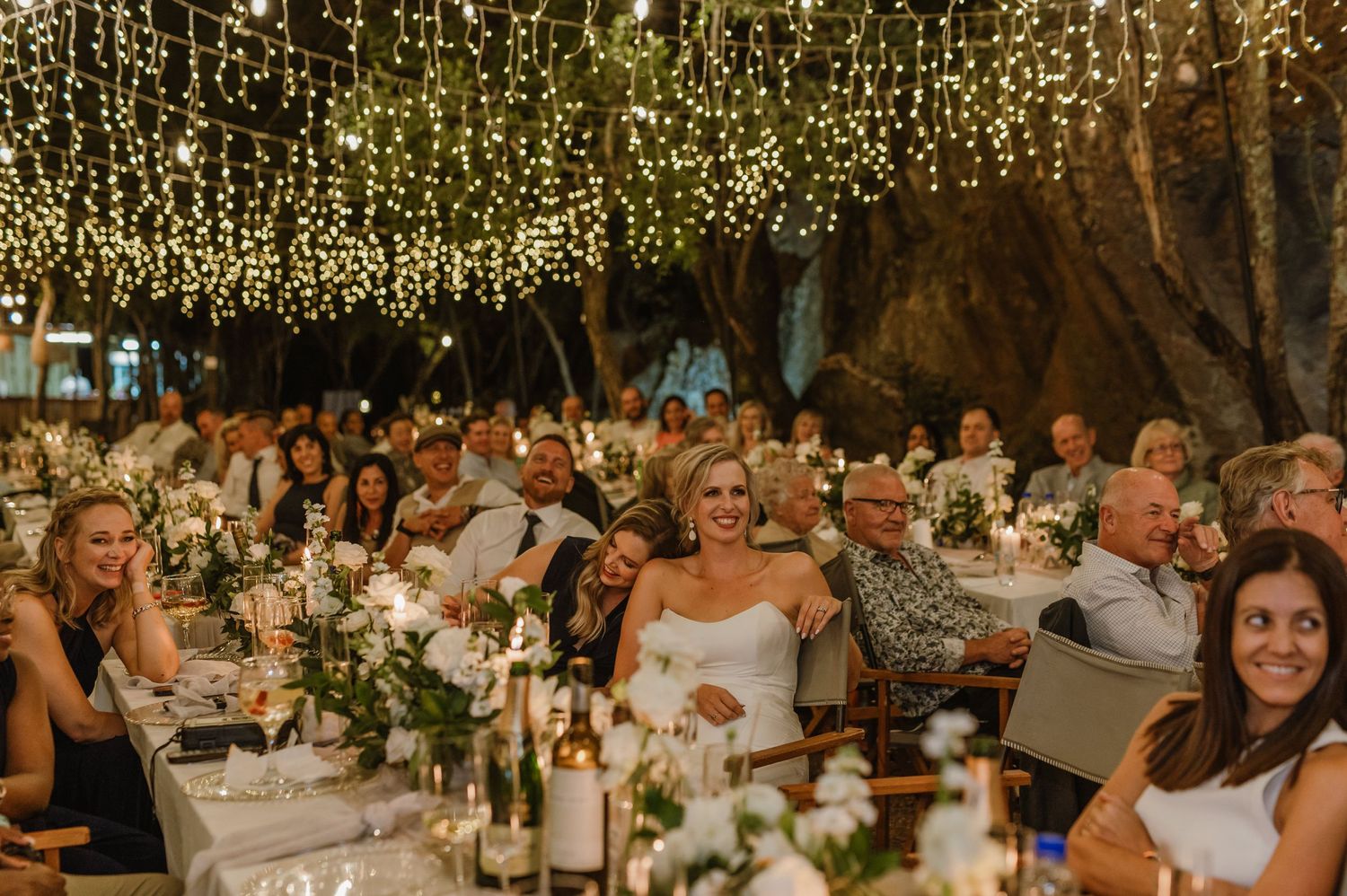 Wedding guests seated at decorated tables beneath a canopy of warm glowing lights.