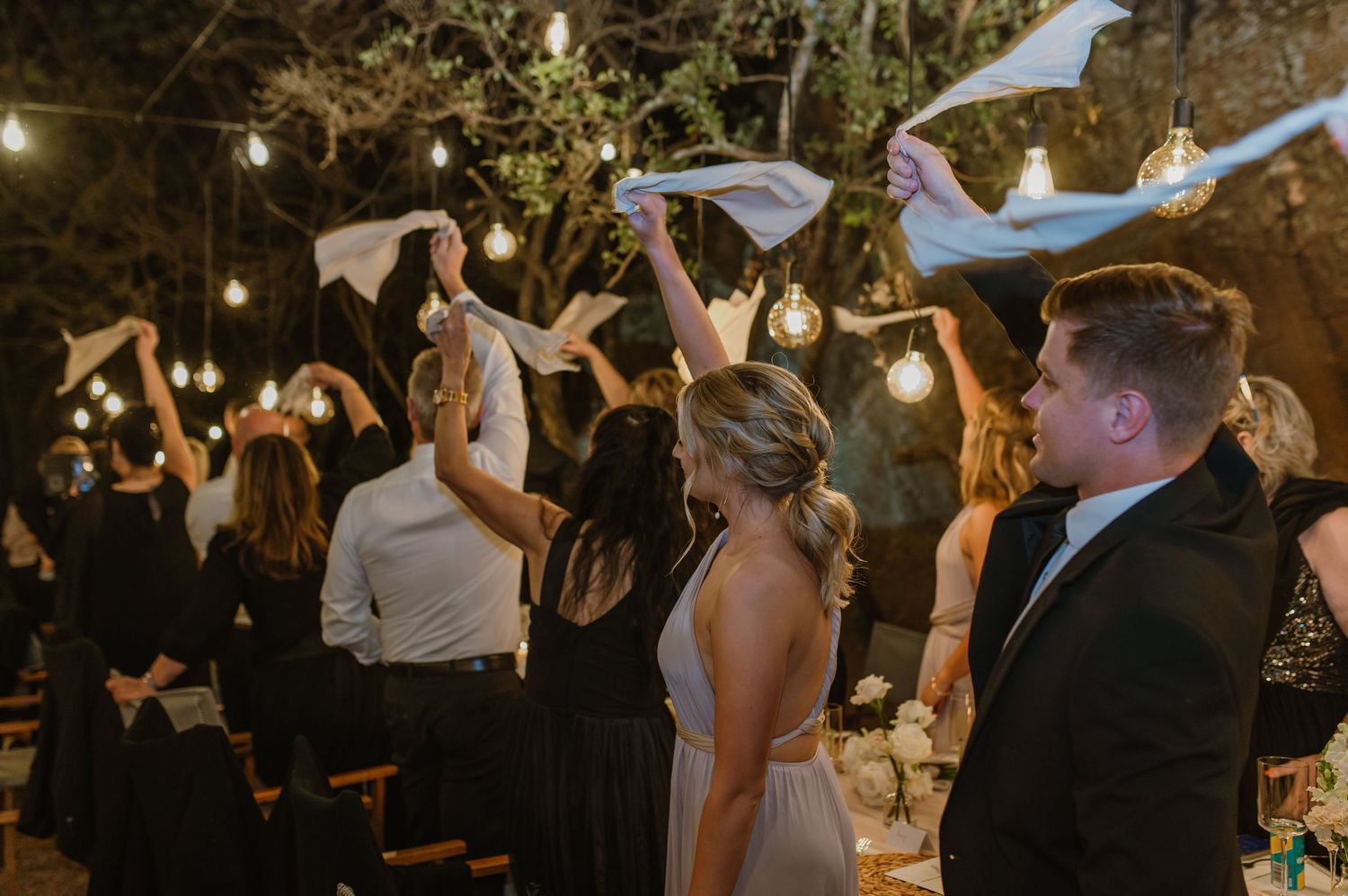 Wedding guests waving napkins during celebration under string lights.