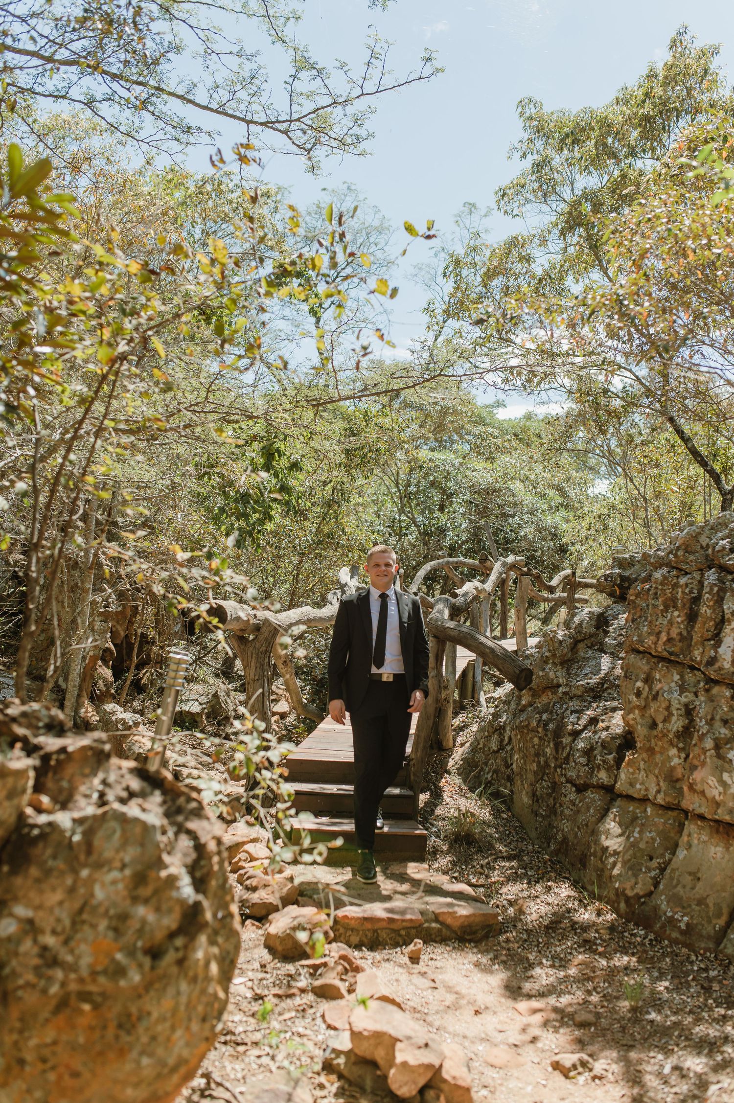 Groom in black suit walking down stone steps surrounded by lush trees at an outdoor wedding venue.