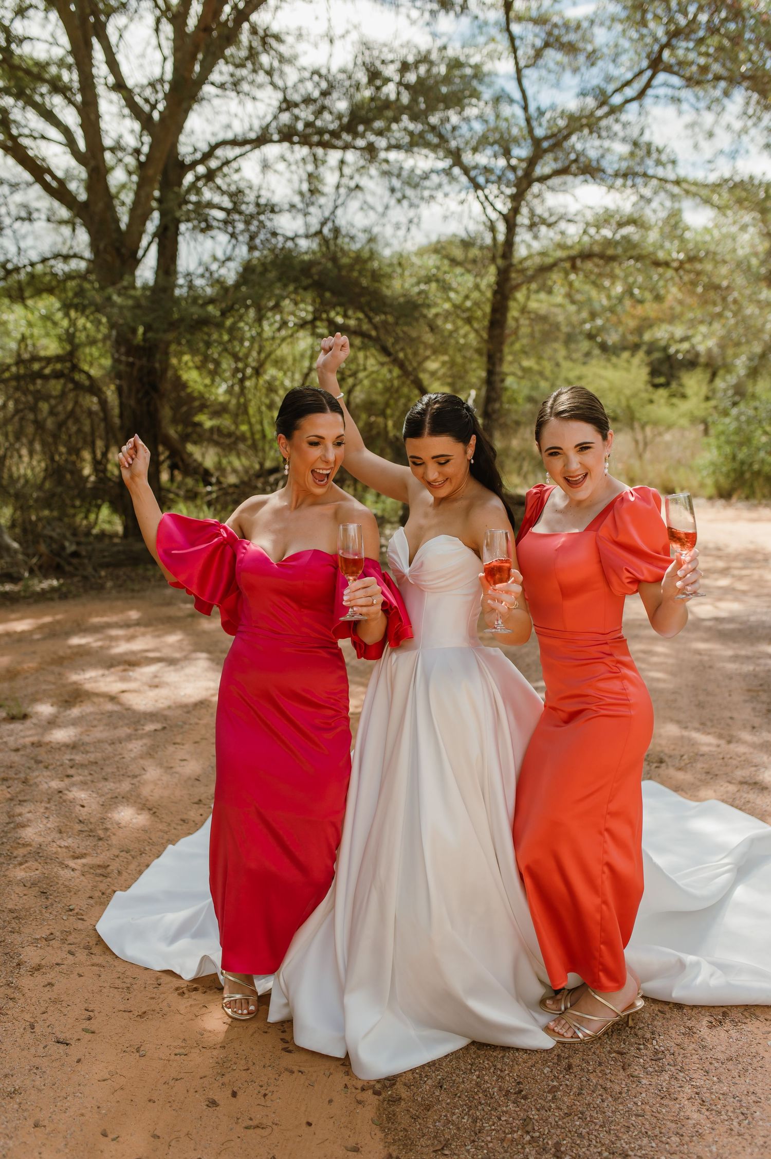 Bride in white gown celebrating with two bridesmaids in red and coral dresses holding champagne glasses outdoors.