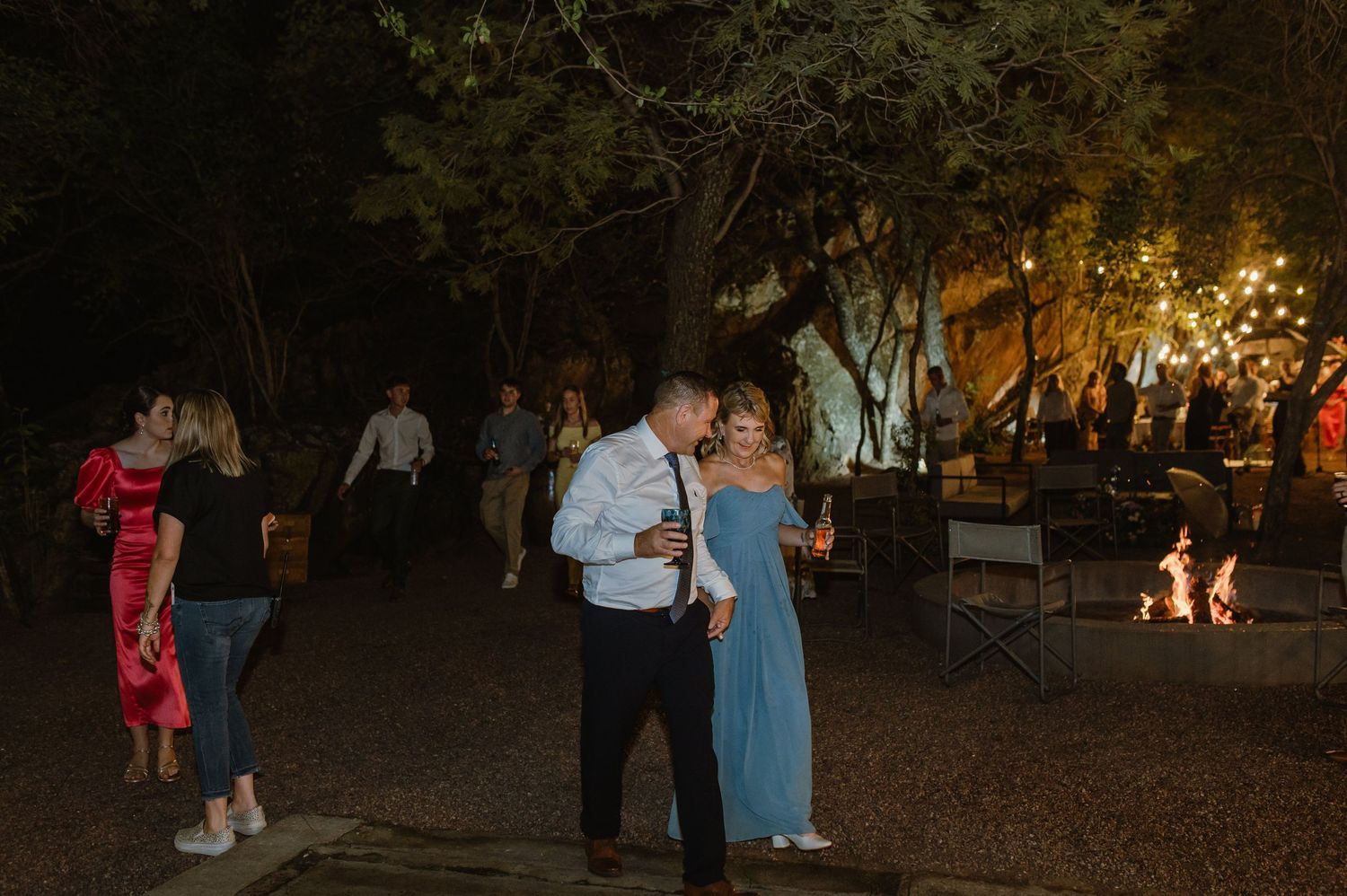 Couple dances outdoors at night wedding reception under string lights surrounded by guests and fire pit.