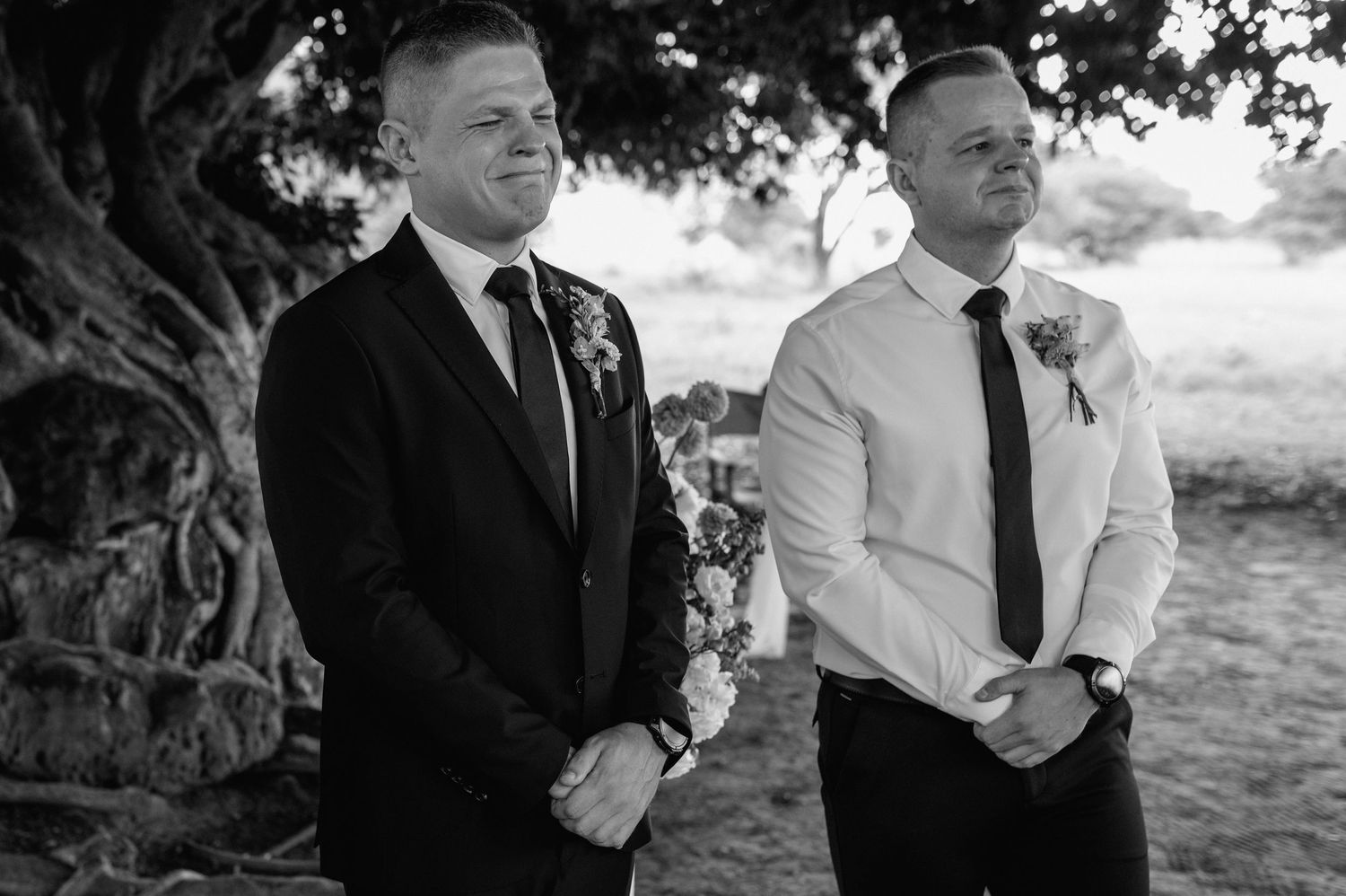 Black and white photo of groom and groomsman wearing boutonnieres standing under large tree during outdoor ceremony.