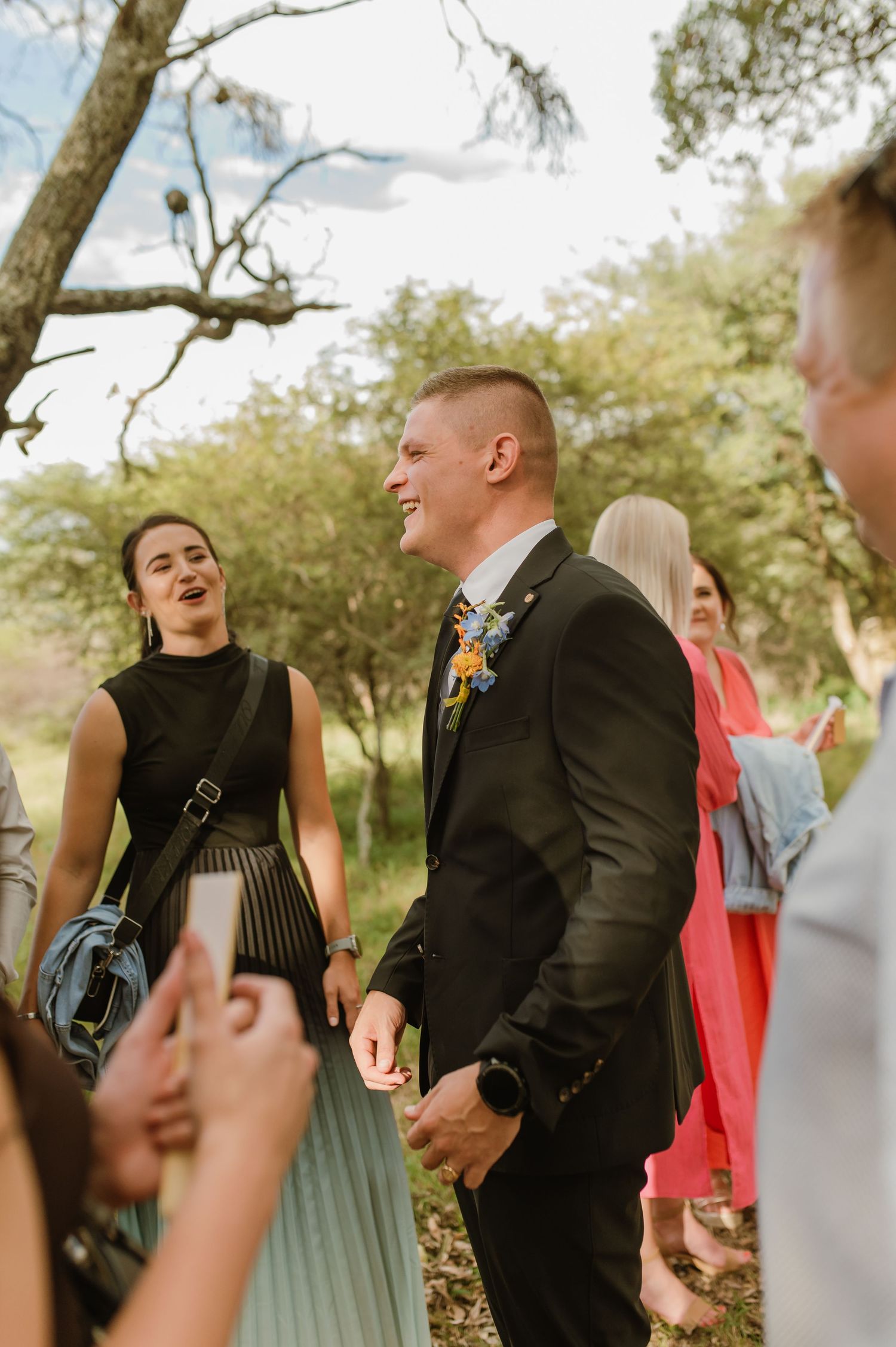 Smiling groom in black suit with floral boutonniere mingling with wedding guests outdoors.