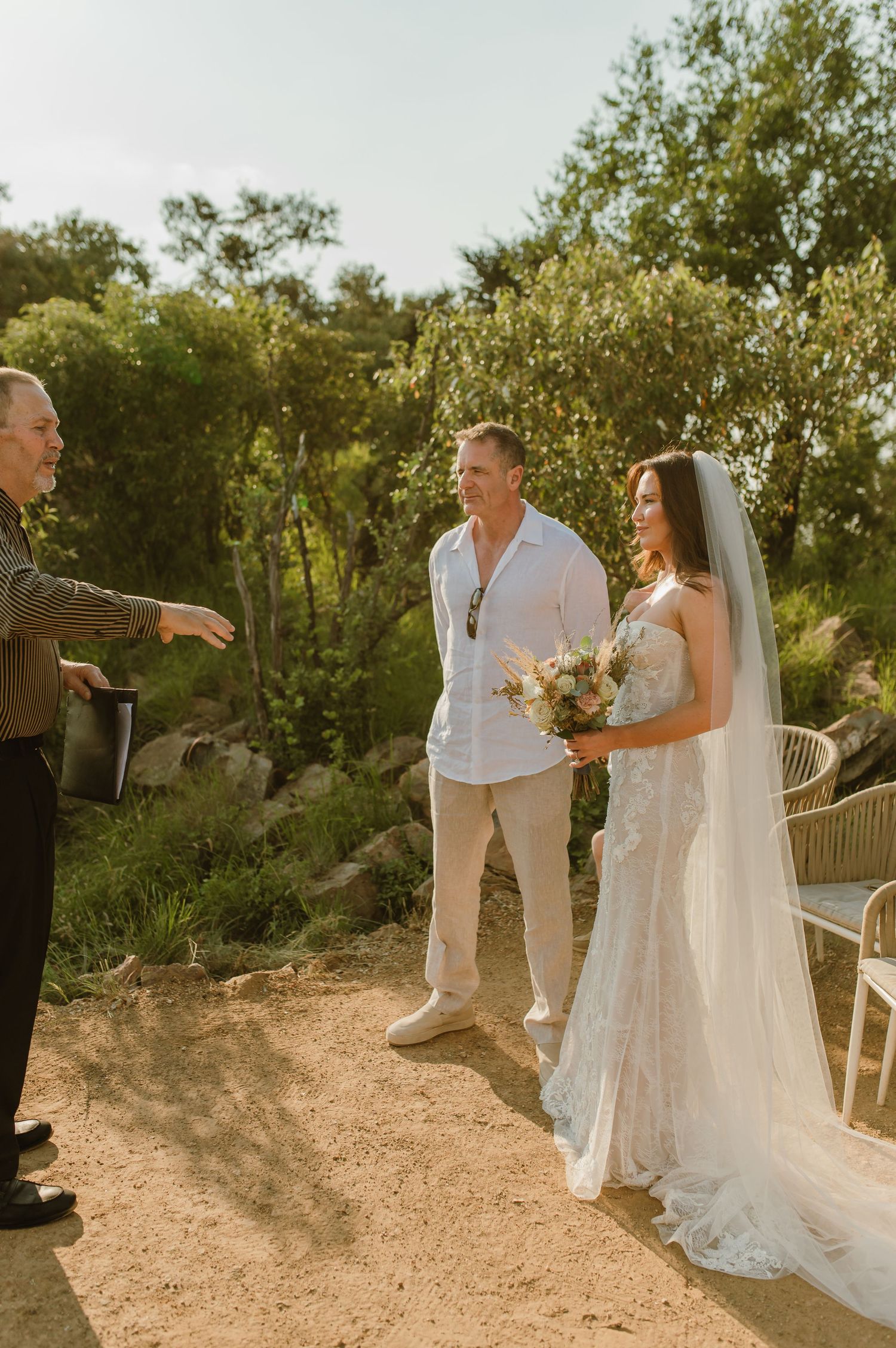 Bride and groom stand with officiant during outdoor wedding ceremony surrounded by lush greenery in natural setting.