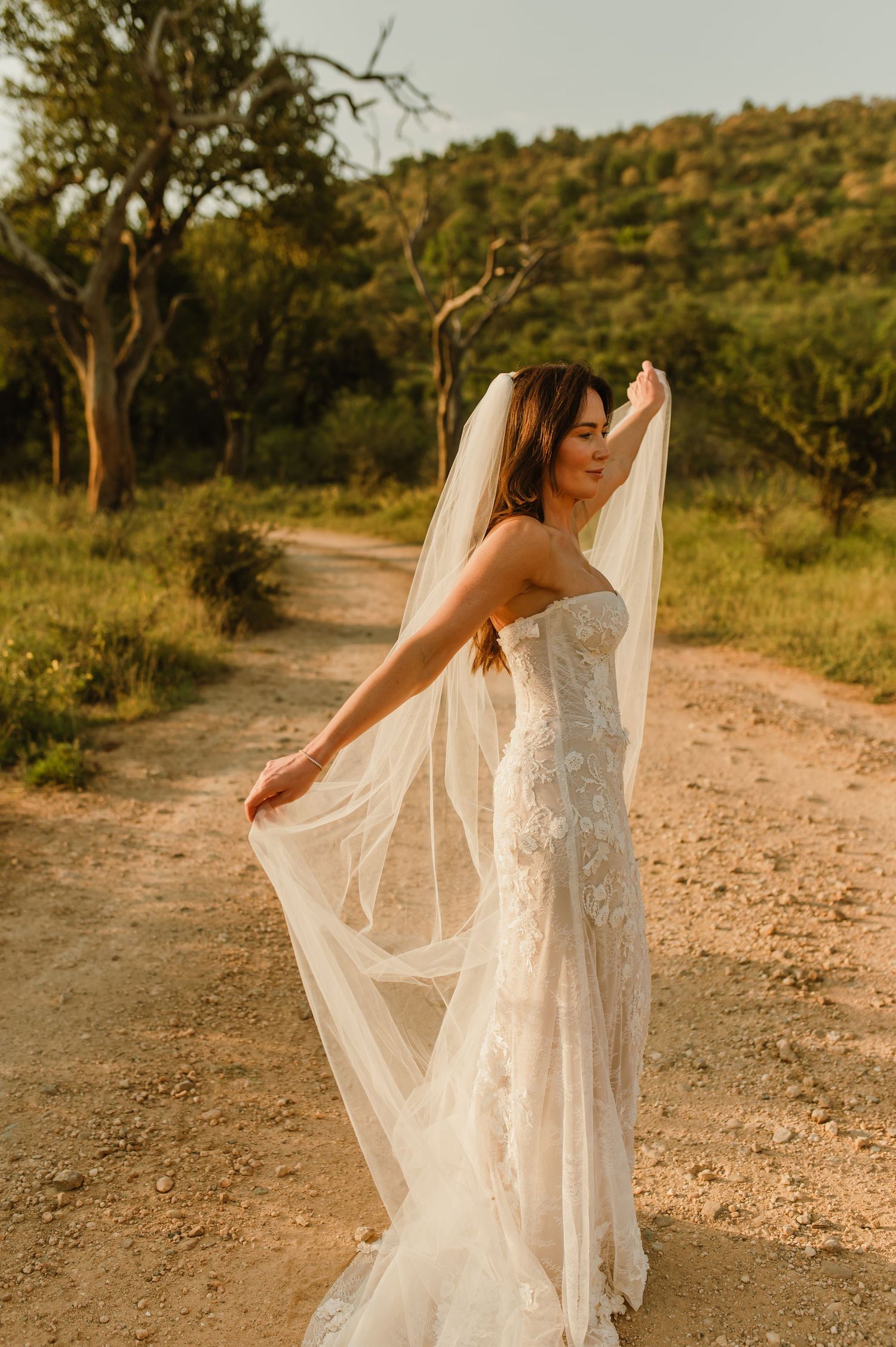 Bride in lace strapless gown holds flowing veil on rustic dirt path at golden hour.