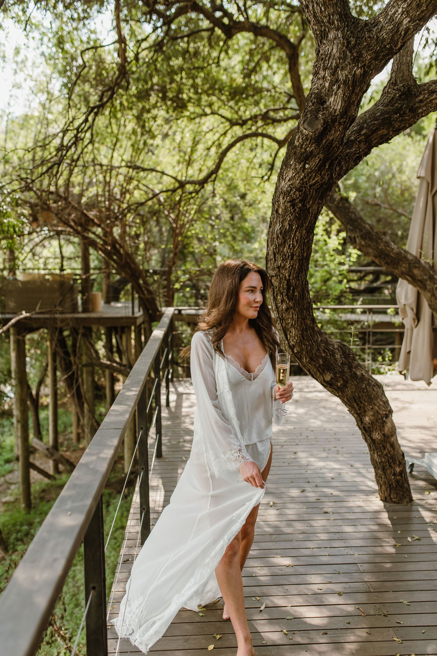 Woman in white flowing dress holding champagne on a wooden walkway surrounded by trees.