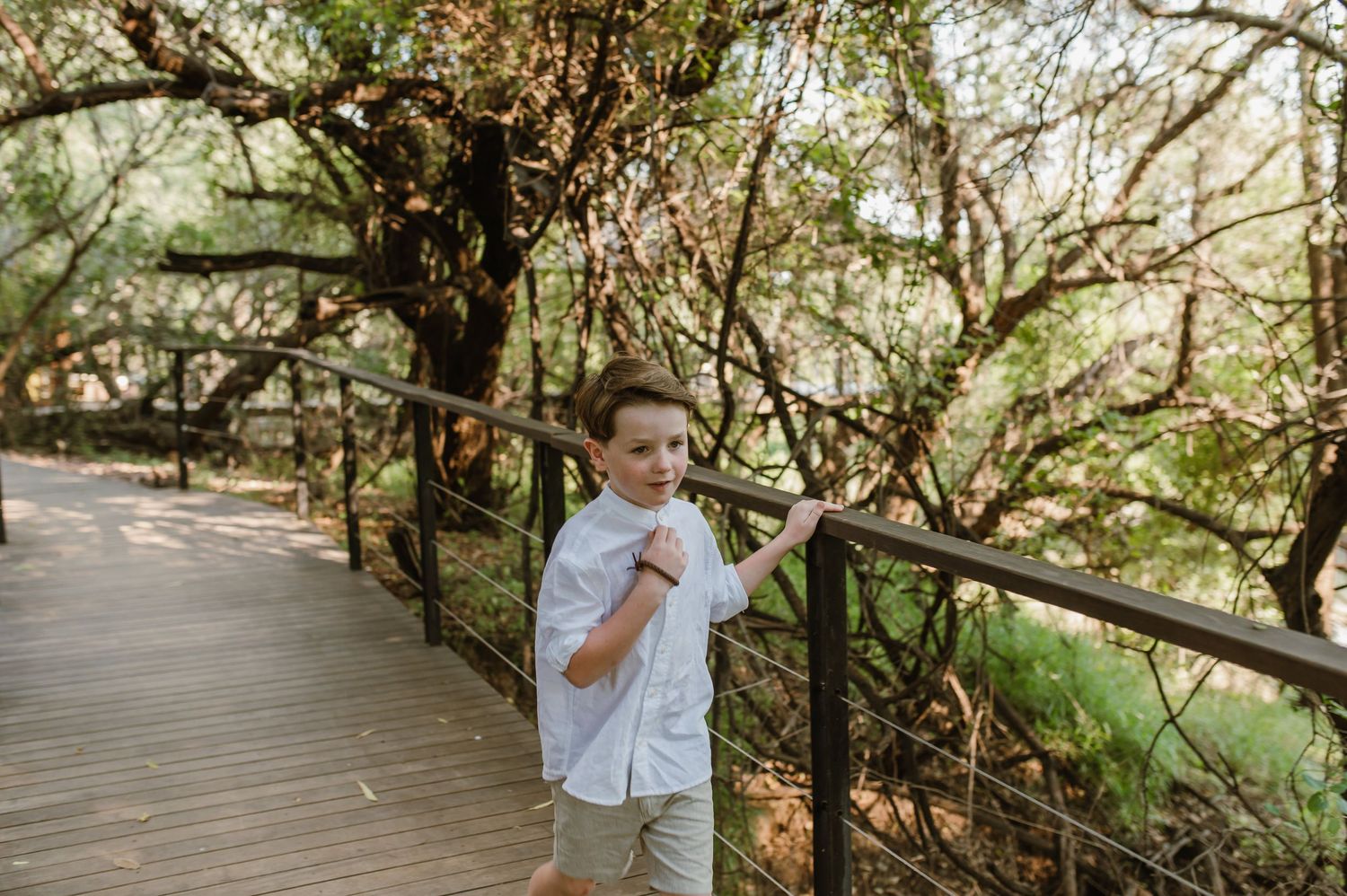 A young boy in a white shirt stands on a wooden boardwalk surrounded by lush trees.