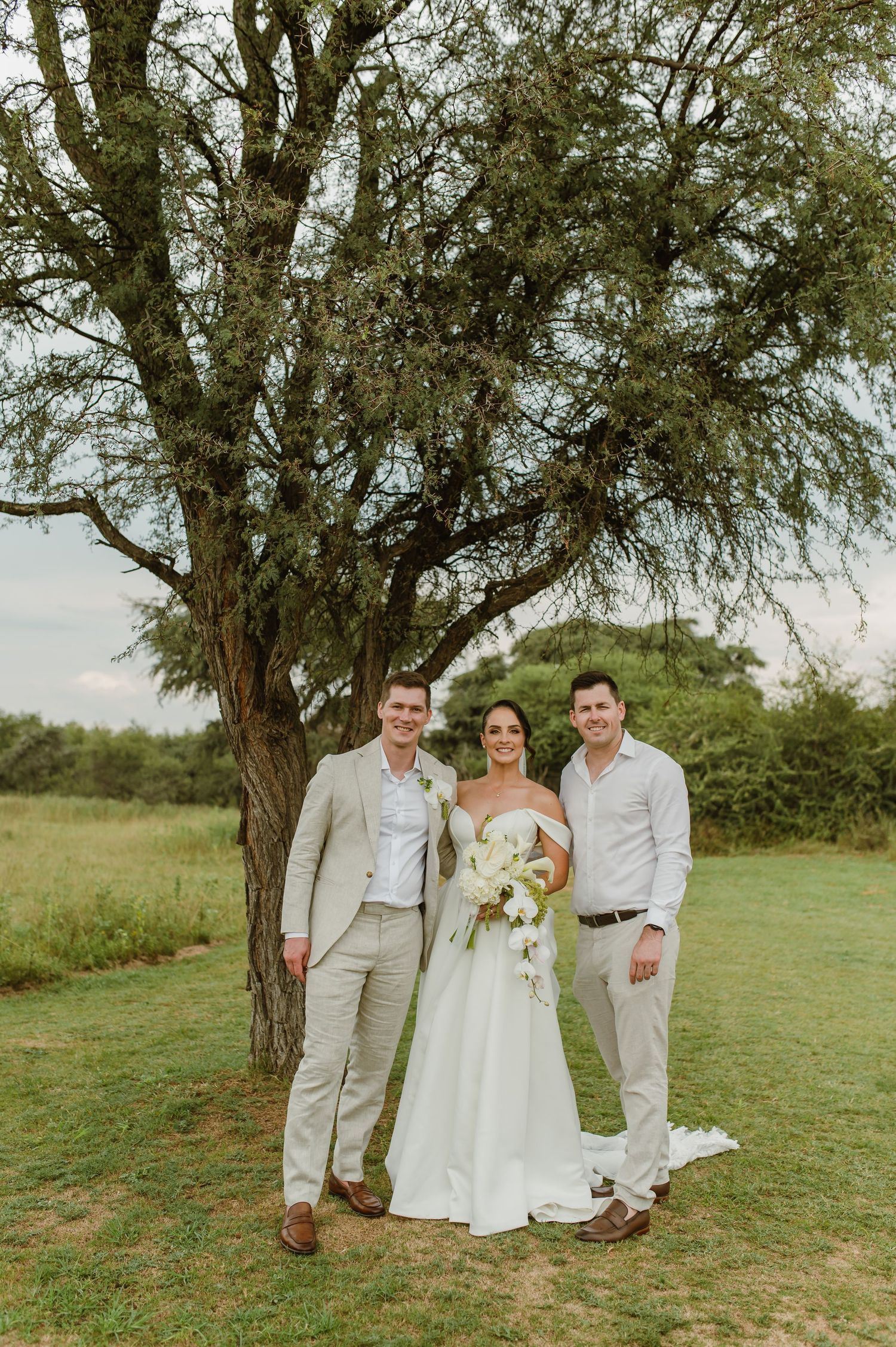A wedding photo taken outdoors under a large tree on a grassy field during a sunny day.