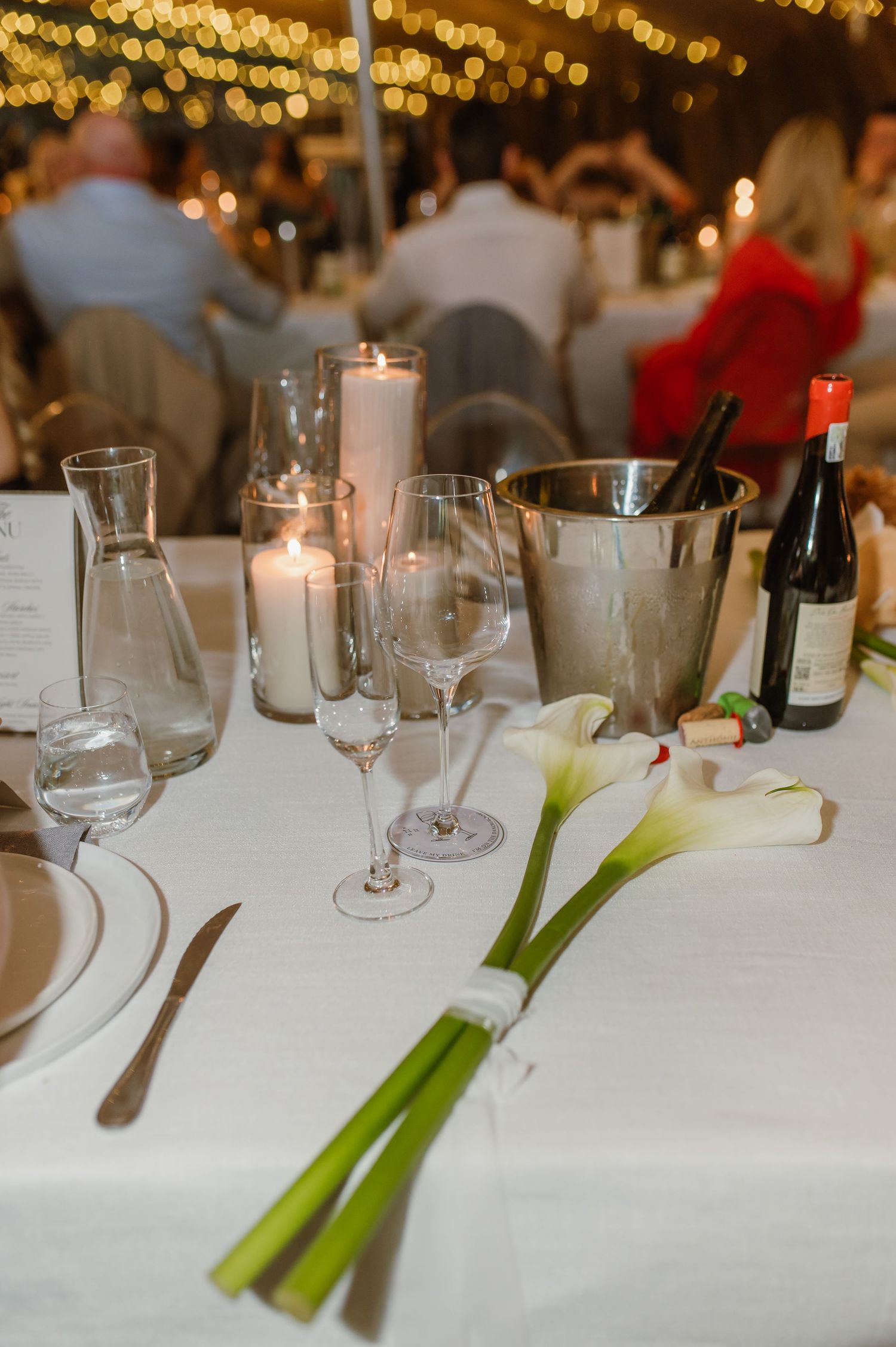 Elegant wedding table setting with white candles, wine glasses, and calla lilies on a white tablecloth under warm lighting.