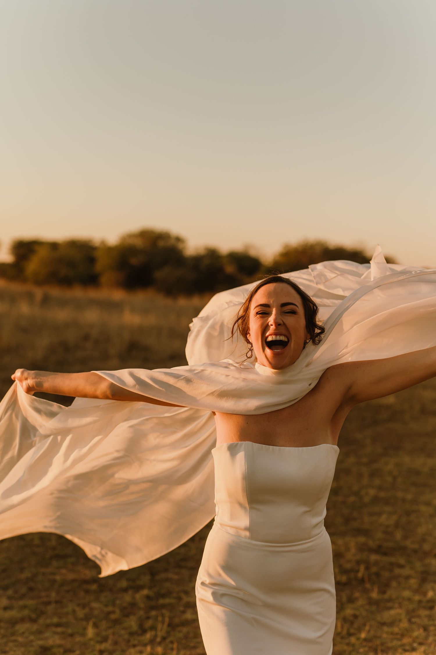 Joyful bride in white strapless gown laughing with veil flowing in golden sunset light in an open field.