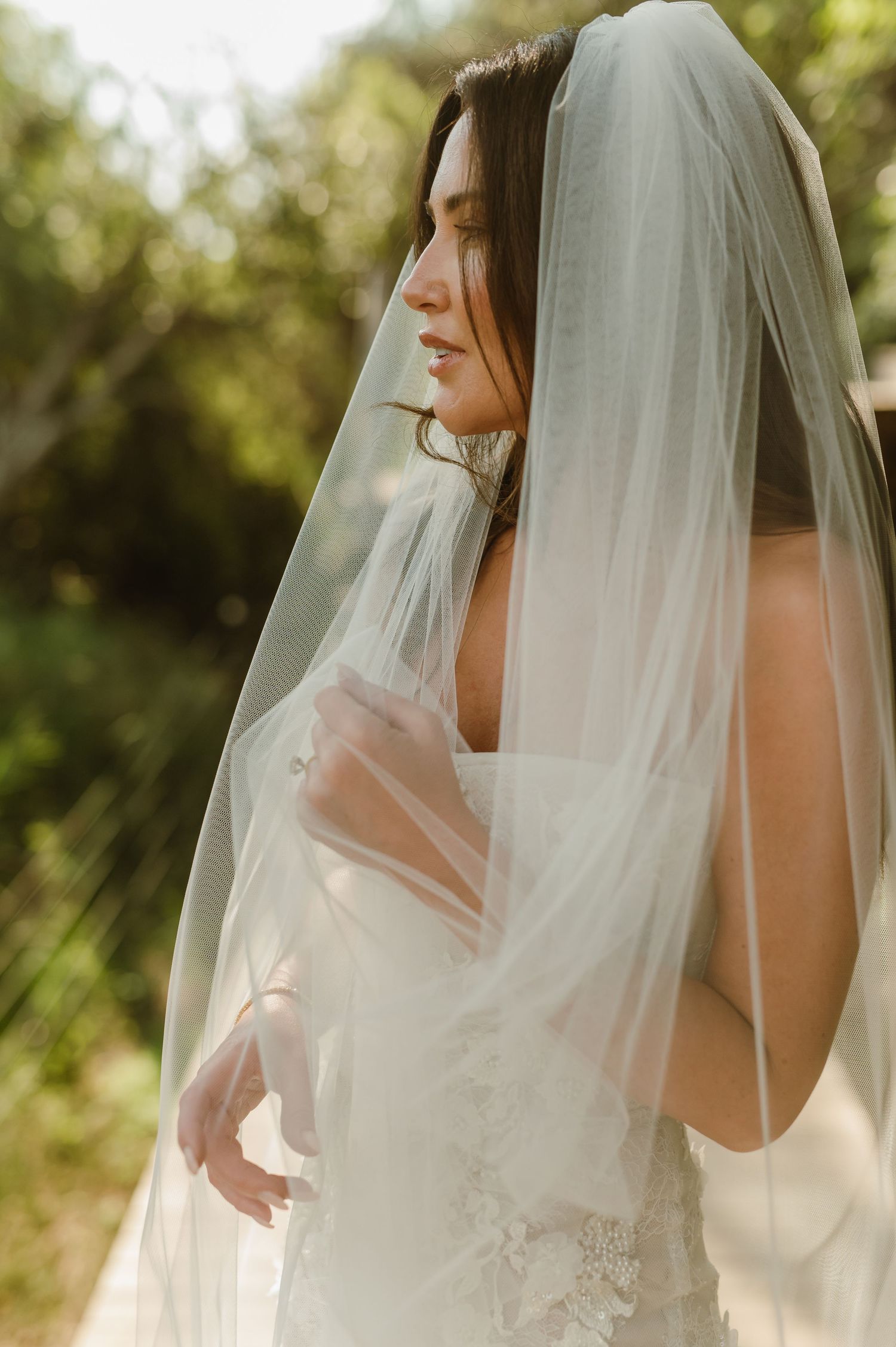 Bride in lace wedding dress with sheer veil draped over face in sunlit garden setting.