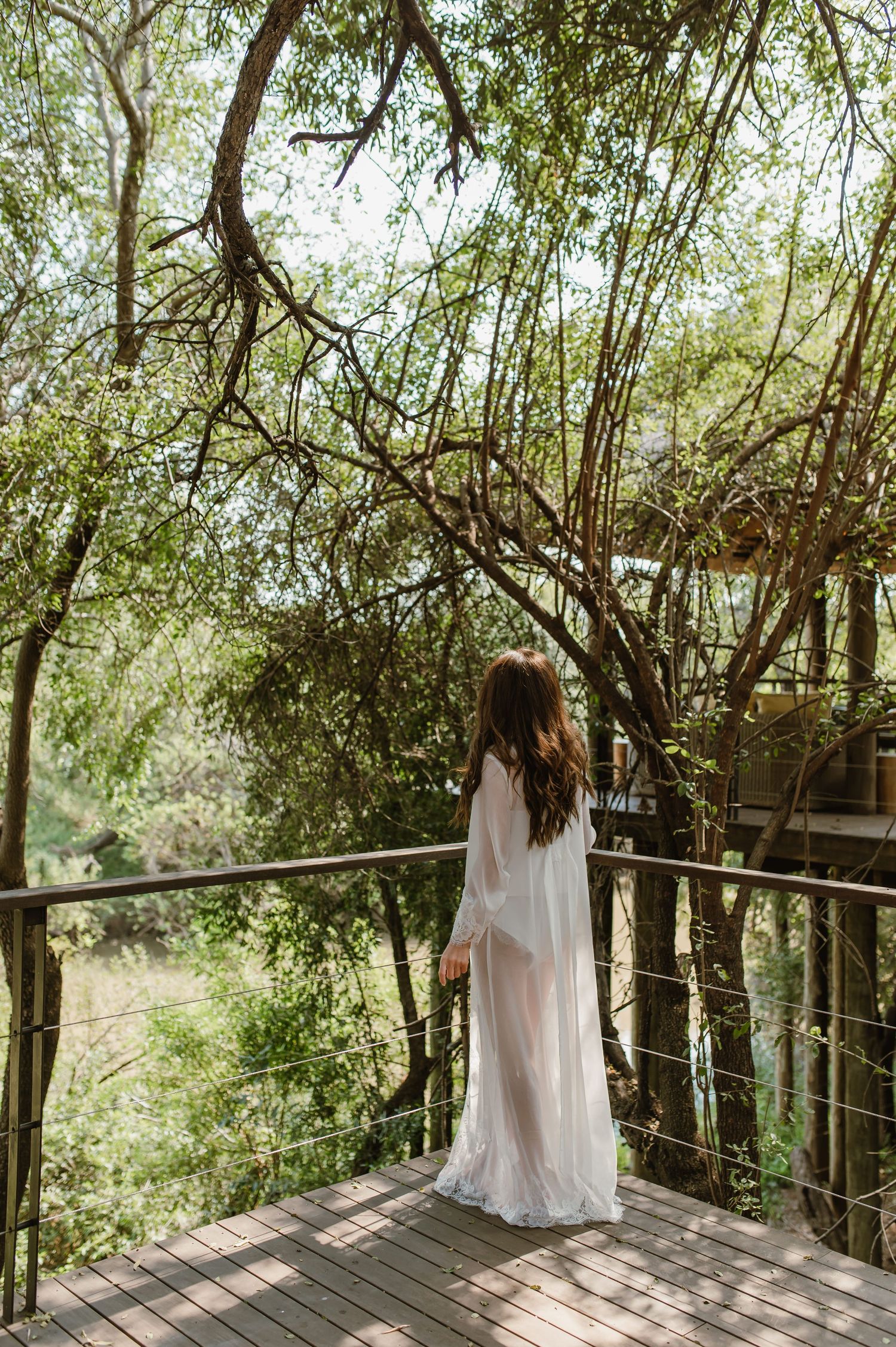 Bride in a flowing white sheer robe standing on a wooden deck surrounded by trees.