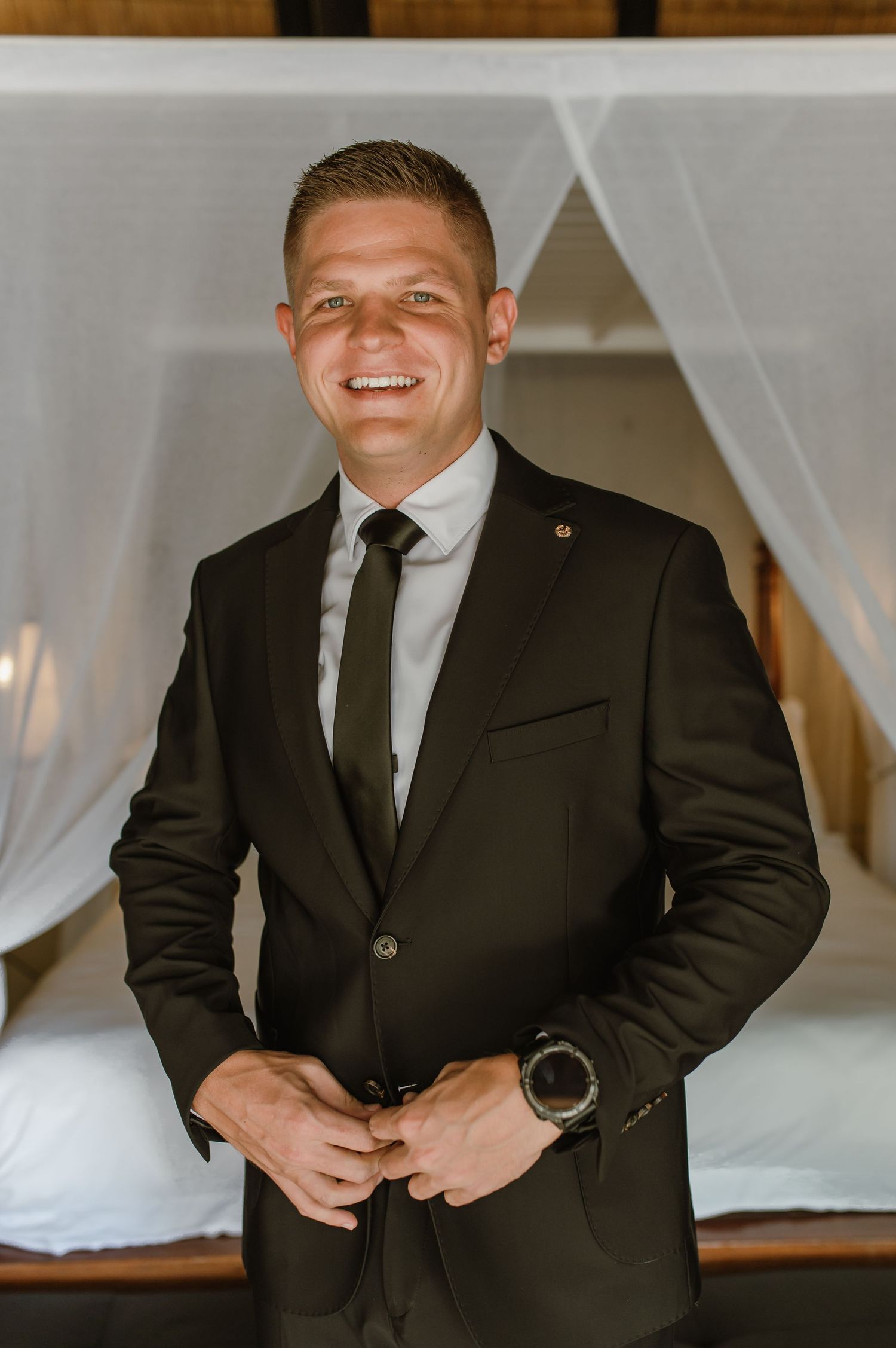 Smiling groom in black suit and tie buttoning jacket in elegant canopy bed suite.
