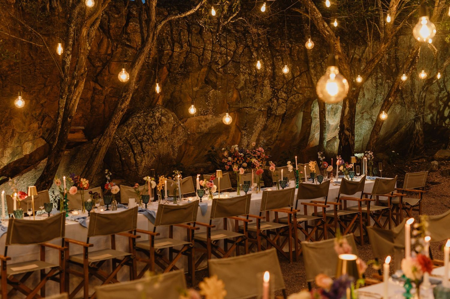 Magical cave wedding reception with long dining tables, Edison bulb string lights, and lush trees.