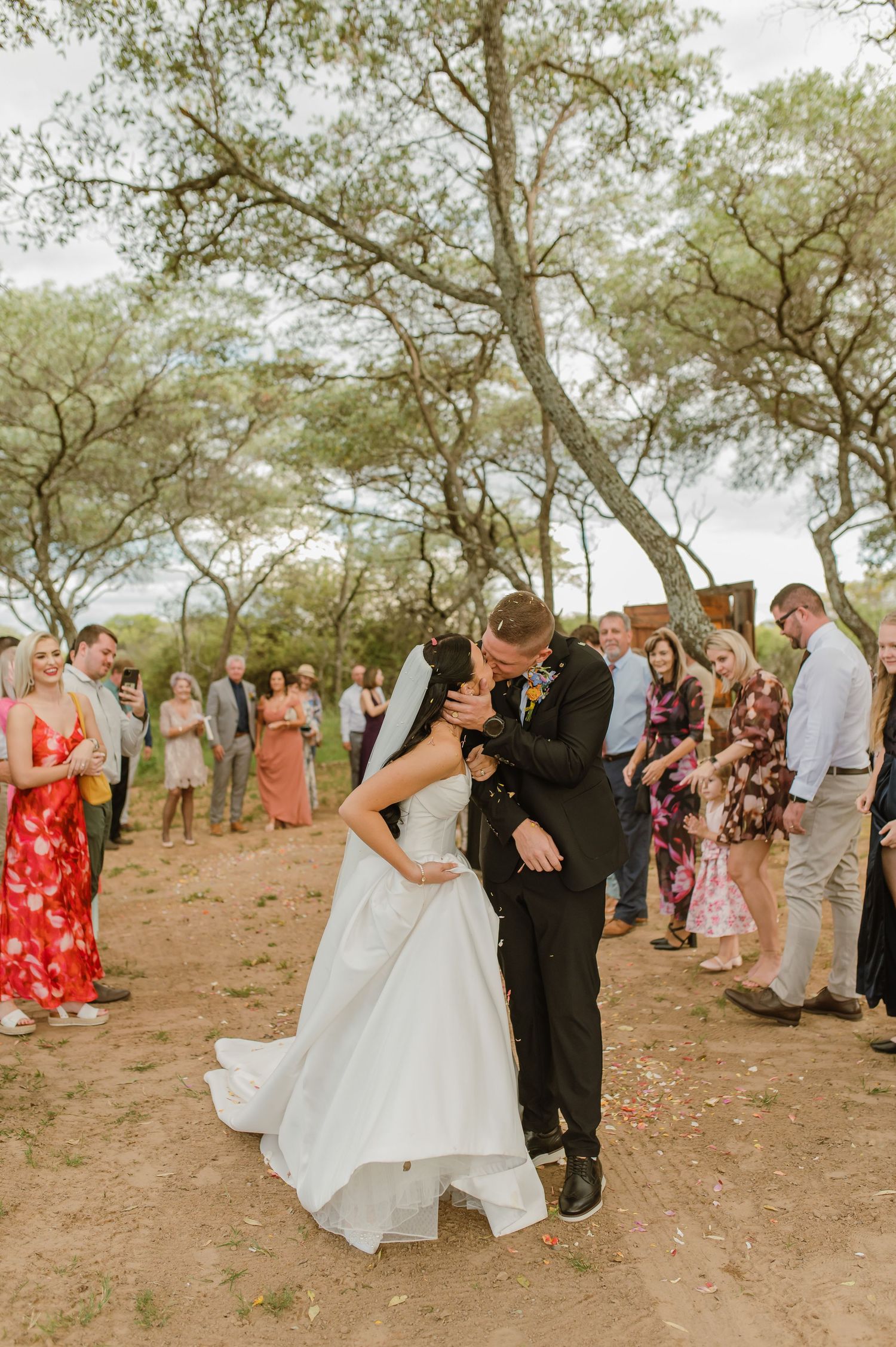 Newlyweds share first kiss as married couple while guests cheer along dirt path at outdoor bush wedding.