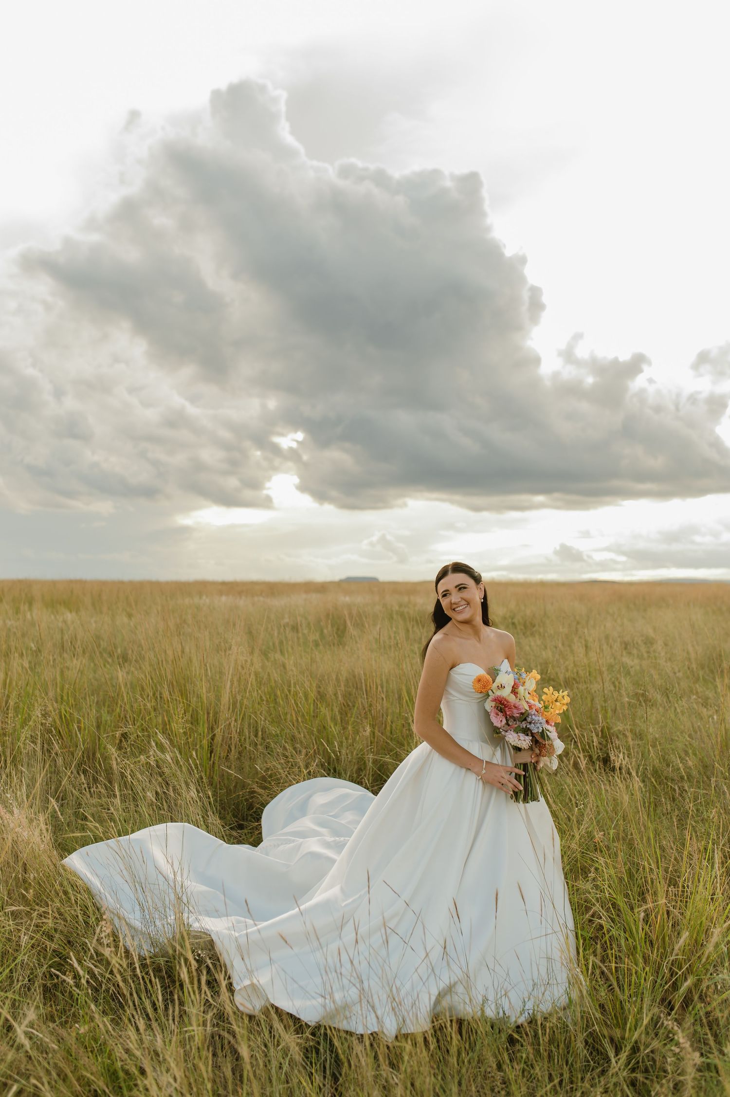 Bride in flowing white strapless gown holds colorful bouquet in golden grass field under dramatic cloudy sky.